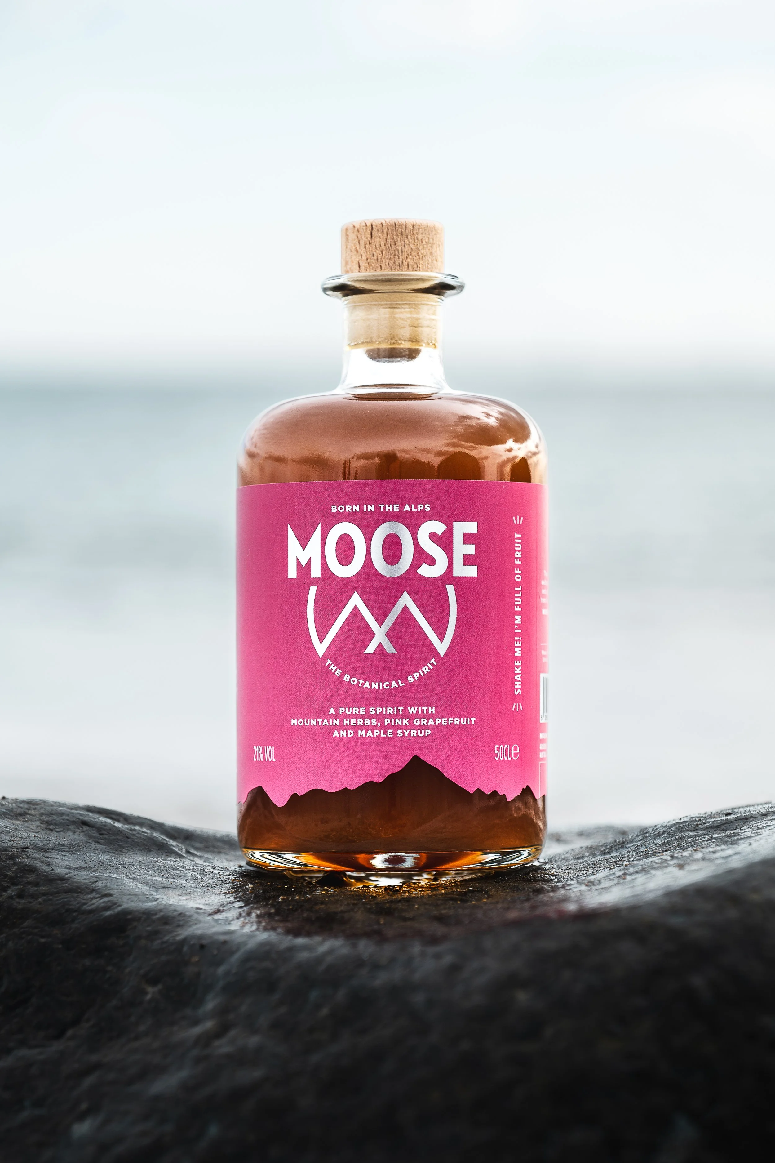 A glass bottle of Moose botanical spirit with a pink label, wooden cap, and mountain illustration, placed on wet dark rock near water.