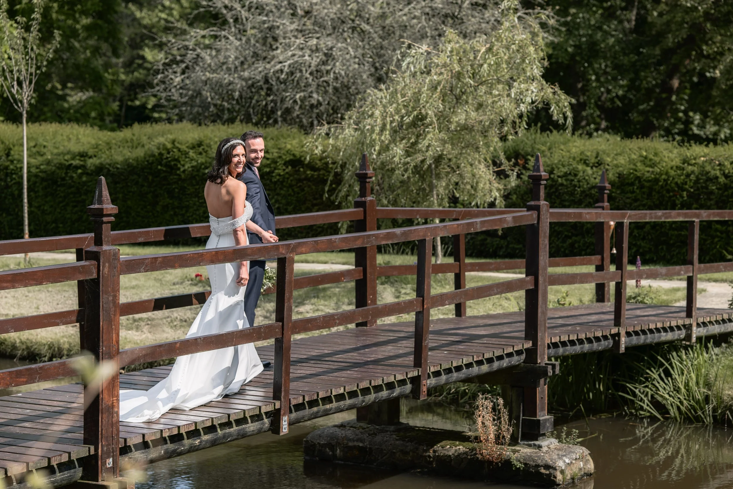 A bride and groom walking on a wooden bridge in a park, smiling and holding hands.