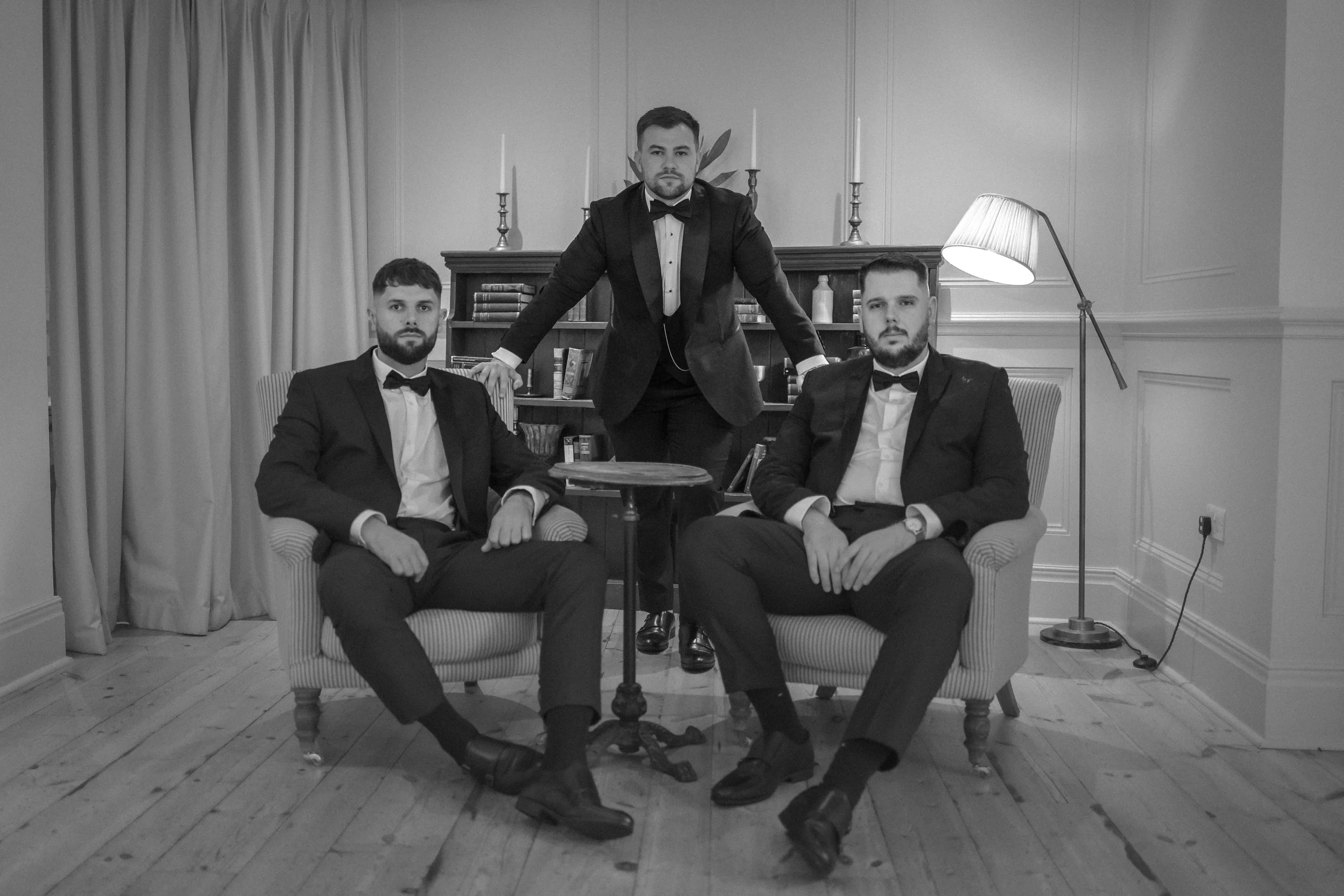 Three men in tuxedos, two seated and one standing behind them in a room with a bookshelf, candlesticks, and a floor lamp, all in black and white.