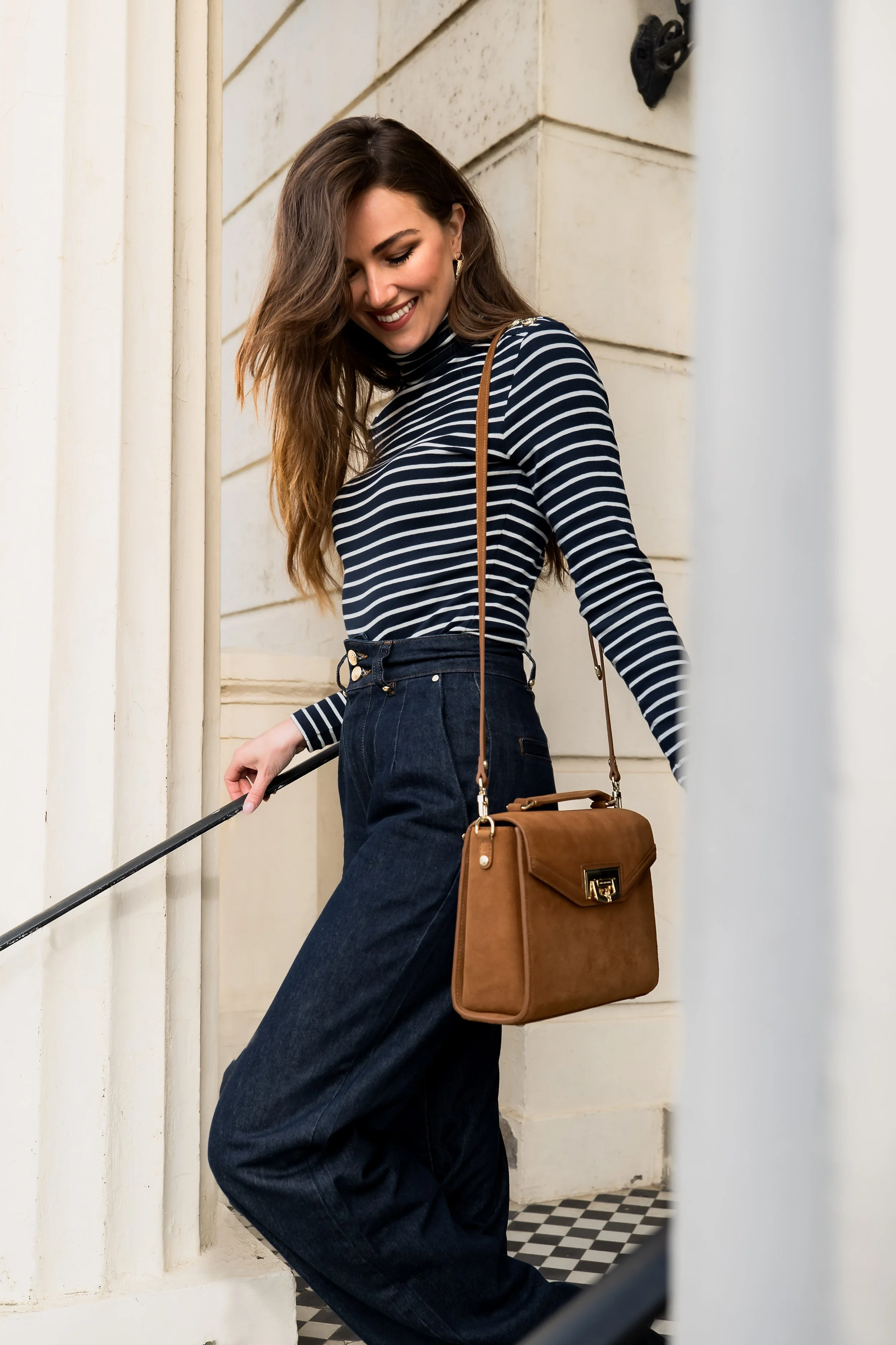 A woman with long brown hair wearing a black and white striped turtleneck and high-waisted dark jeans, smiling and holding a brown handbag, standing near a white building with a black-and-white checkered floor.