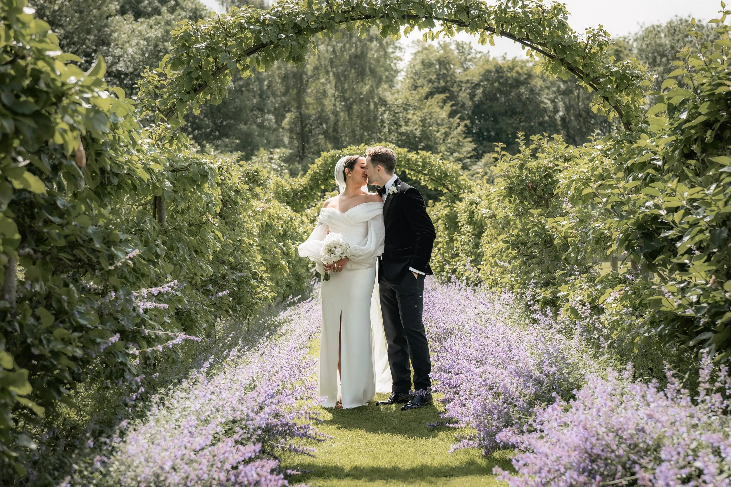 Bride and groom sharing a kiss in a garden with archway and blooming lavender plants.