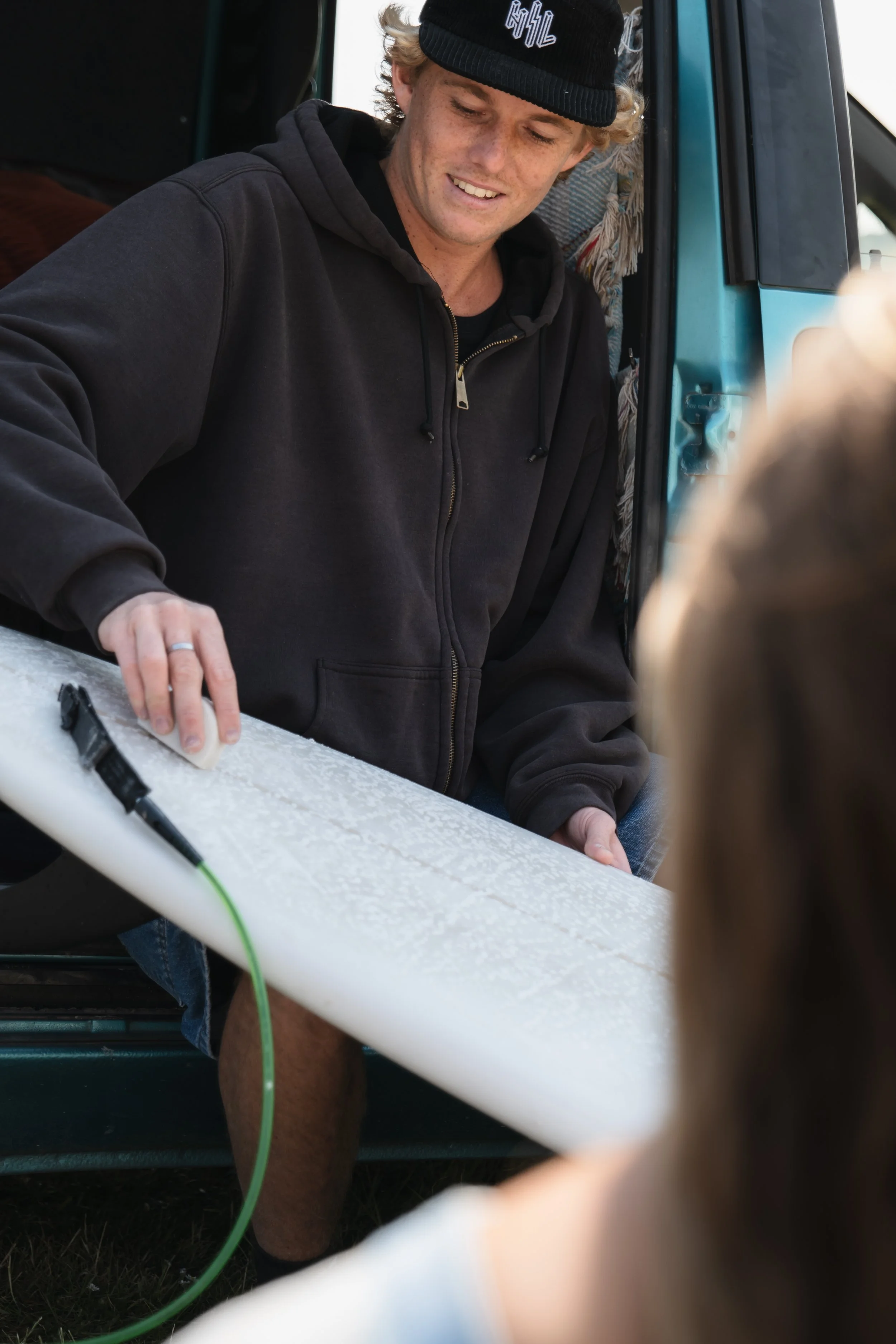 A young man in a black hoodie and beanie smoothing the surface of a surfboard, with a person in the foreground observing.