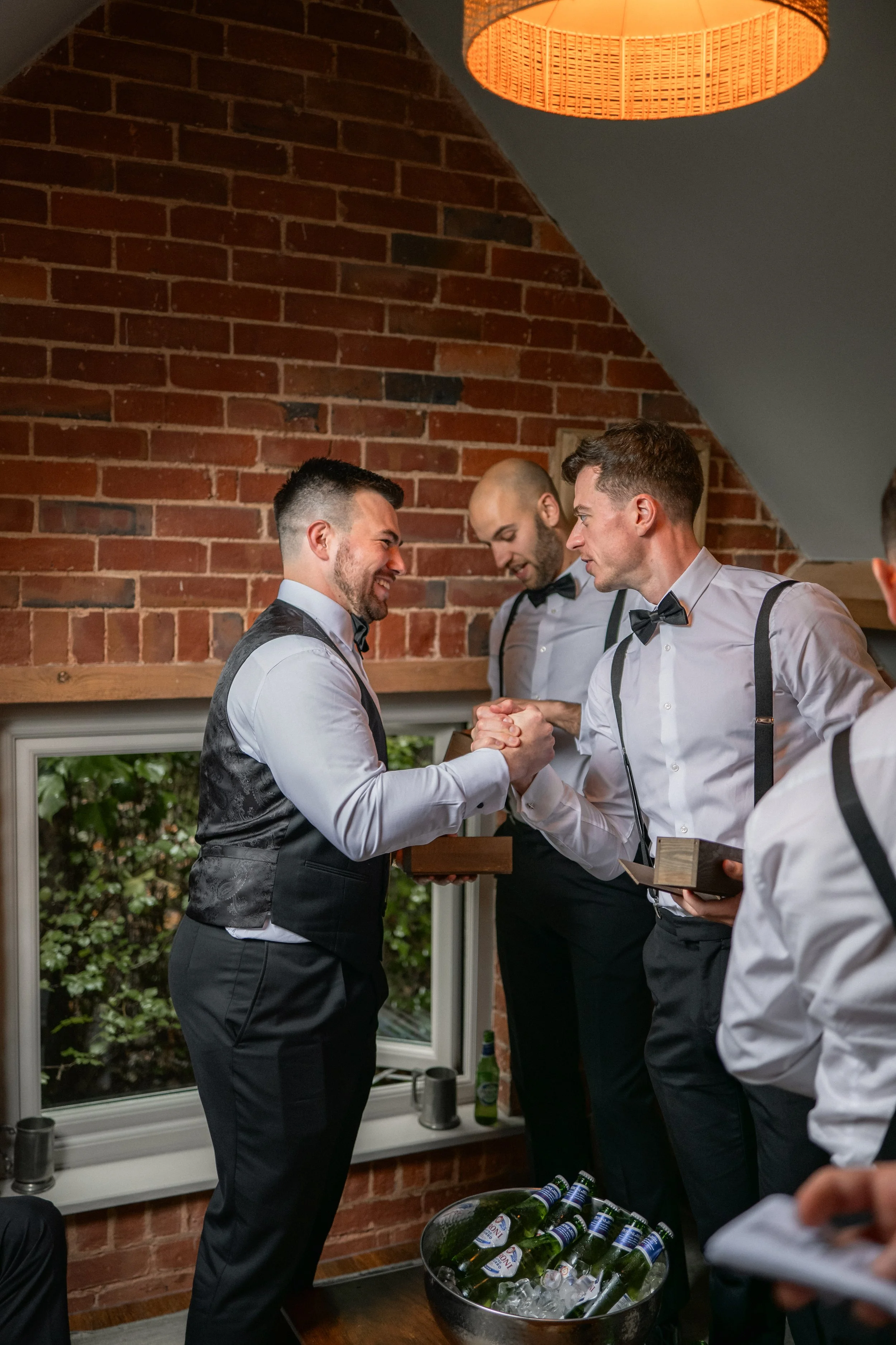 Two men in formal attire shake hands, smiling, during a celebration or wedding; other men in formal wear stand nearby, one with a tray of drinks and another holding a box, in a room with a brick wall and a window showing greenery outside, decorated w