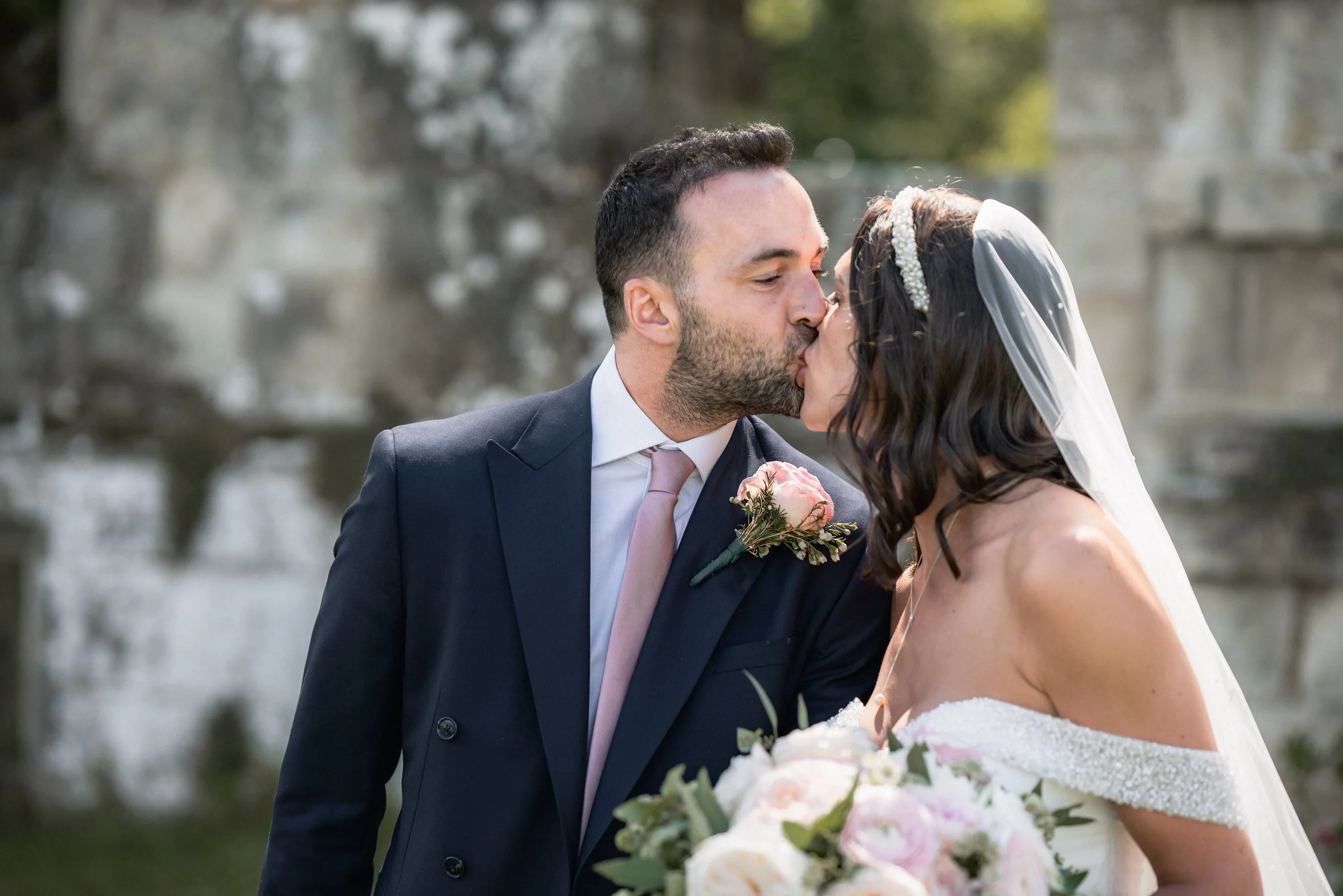 A bride and groom sharing a kiss outdoors, with the groom wearing a dark suit with a pink tie and boutonniere, and the bride wearing a strapless white wedding dress, veil, and holding a bouquet of pink and white flowers.