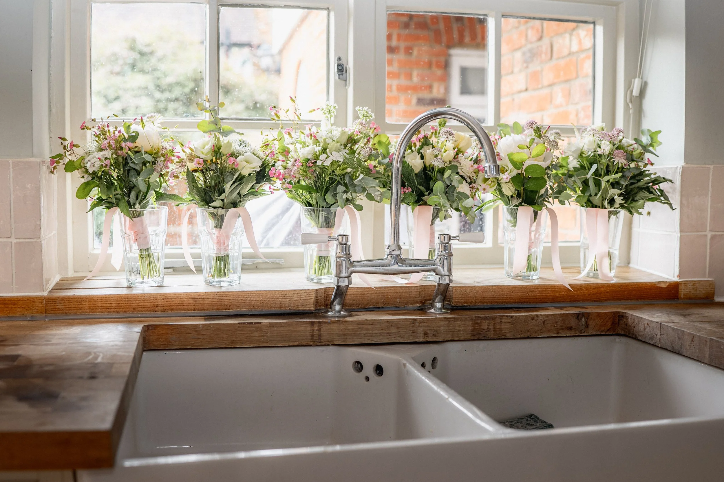 A kitchen window sill decorated with five glass vases filled with pink and white flowers, each tied with a pink ribbon, with a kitchen sink and faucet below.