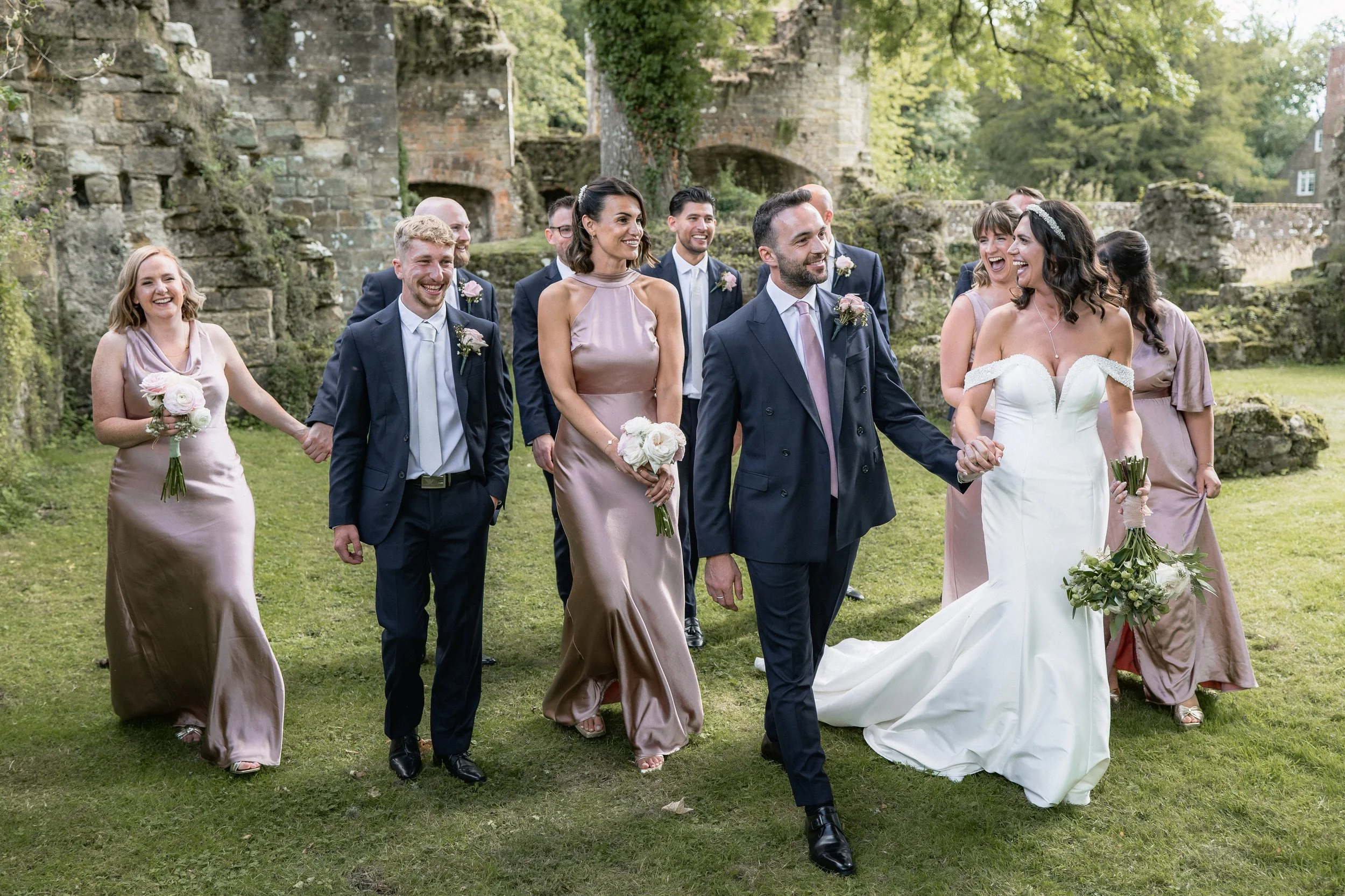 A group of wedding guests walking outdoors on green grass, with a bride in a white gown and groom in a dark suit at the front. The guests are dressed in formal attire, holding bouquets, and smiling under trees and old stone ruins.