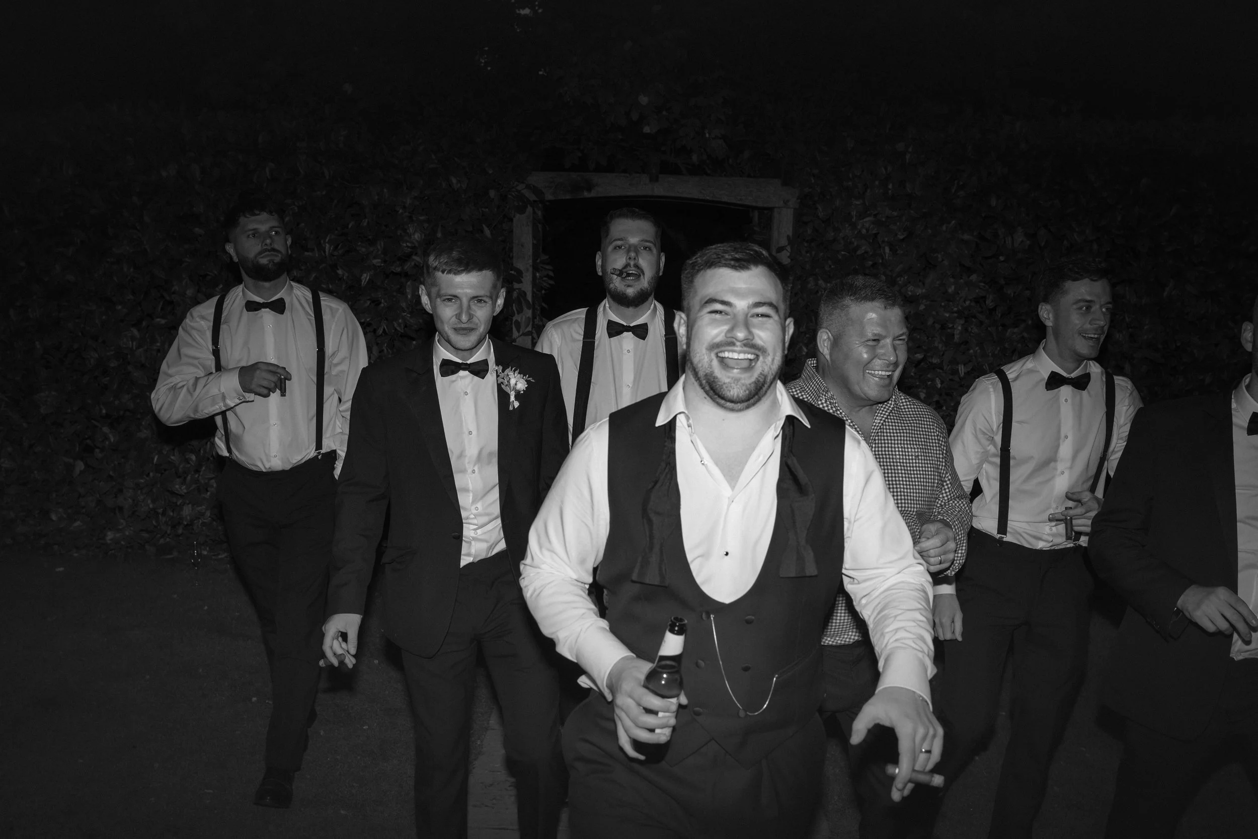 Group of men in formal attire at night, some smiling and holding drinks, outdoors with foliage background.