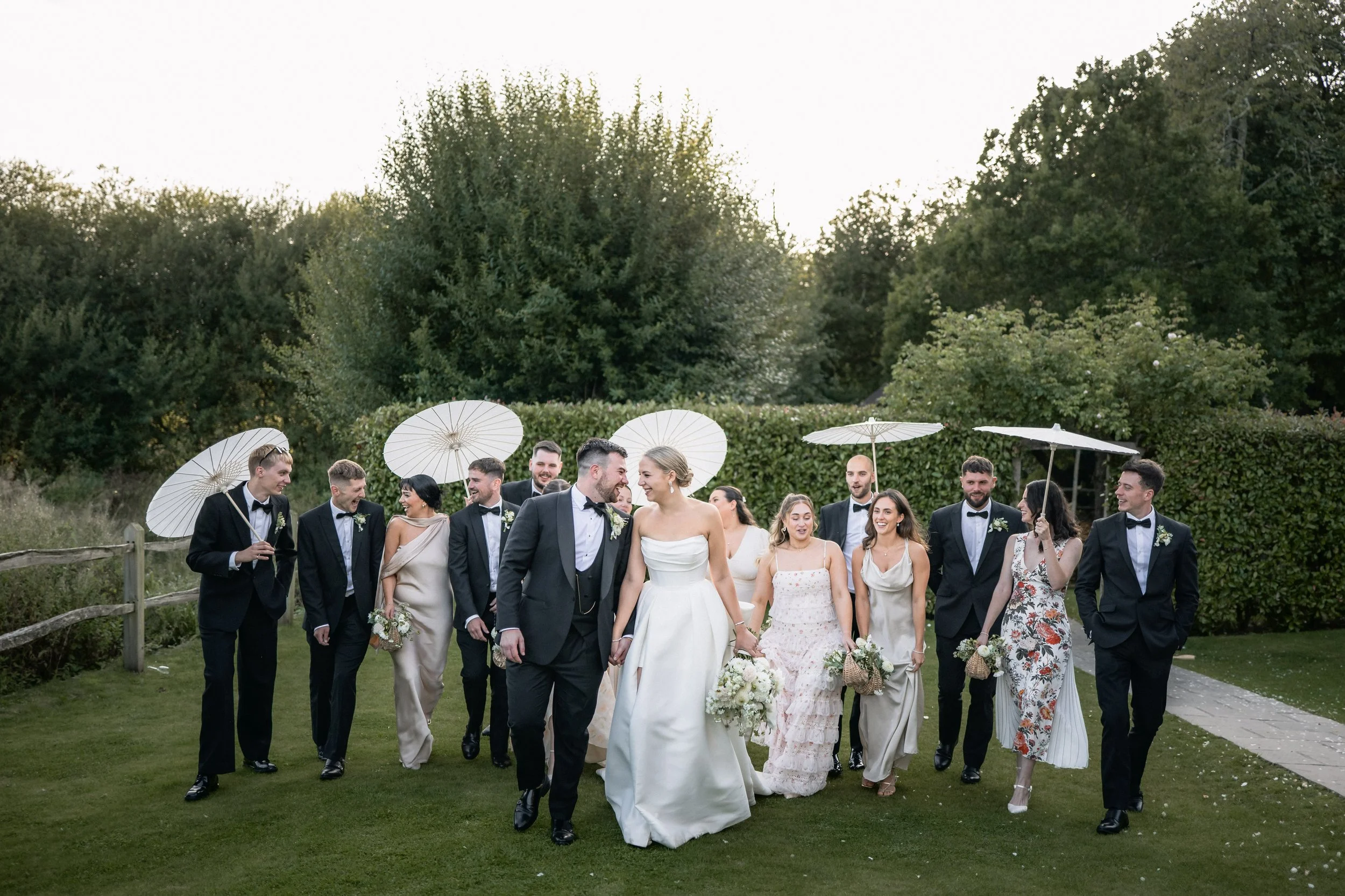A wedding party walking outdoors on grass, with the bride and groom in front, surrounded by bridesmaids and groomsmen holding parasols, with trees in the background.