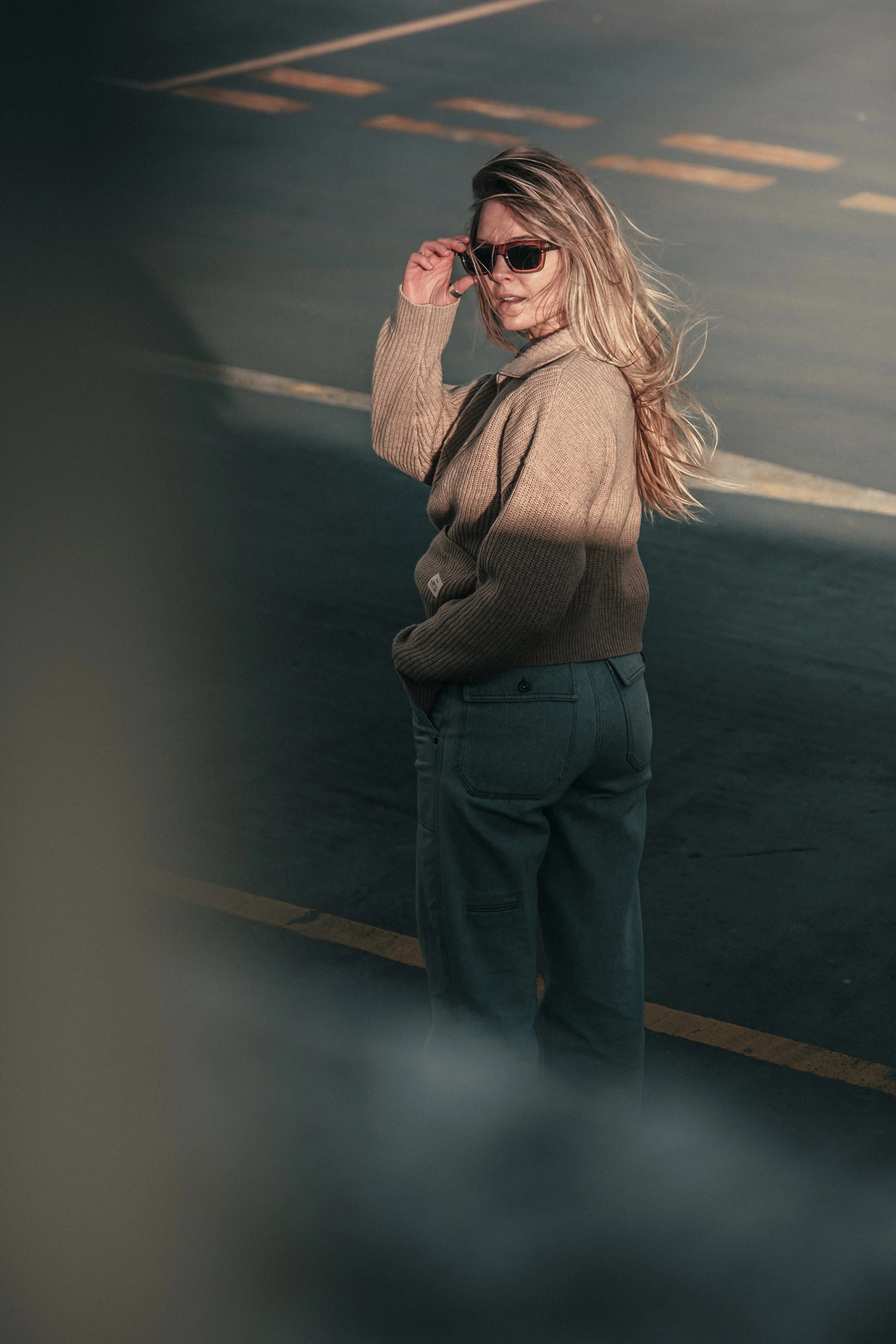 A woman with long blonde hair, wearing sunglasses, a brown sweater, and blue jeans, standing on an asphalt surface, possibly a parking lot.