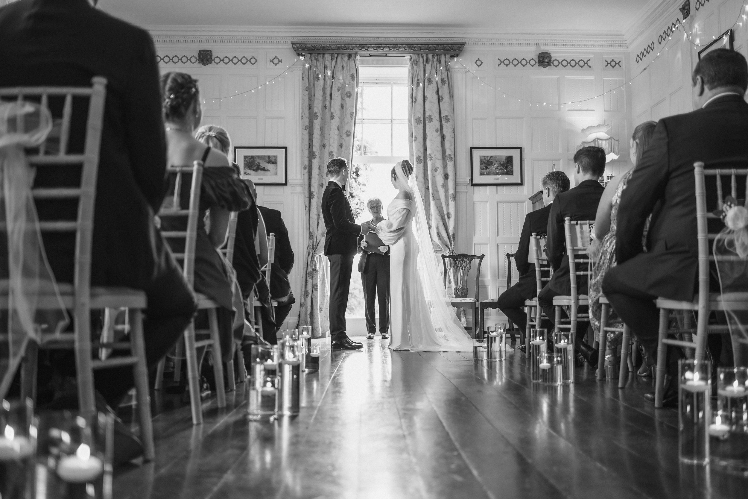 Black and white photo of a wedding ceremony in a decorated room with large window, curtains, and framed pictures. The bride and groom stand facing each other, holding hands, with an officiant between them. Guests sit on either side, facing the couple