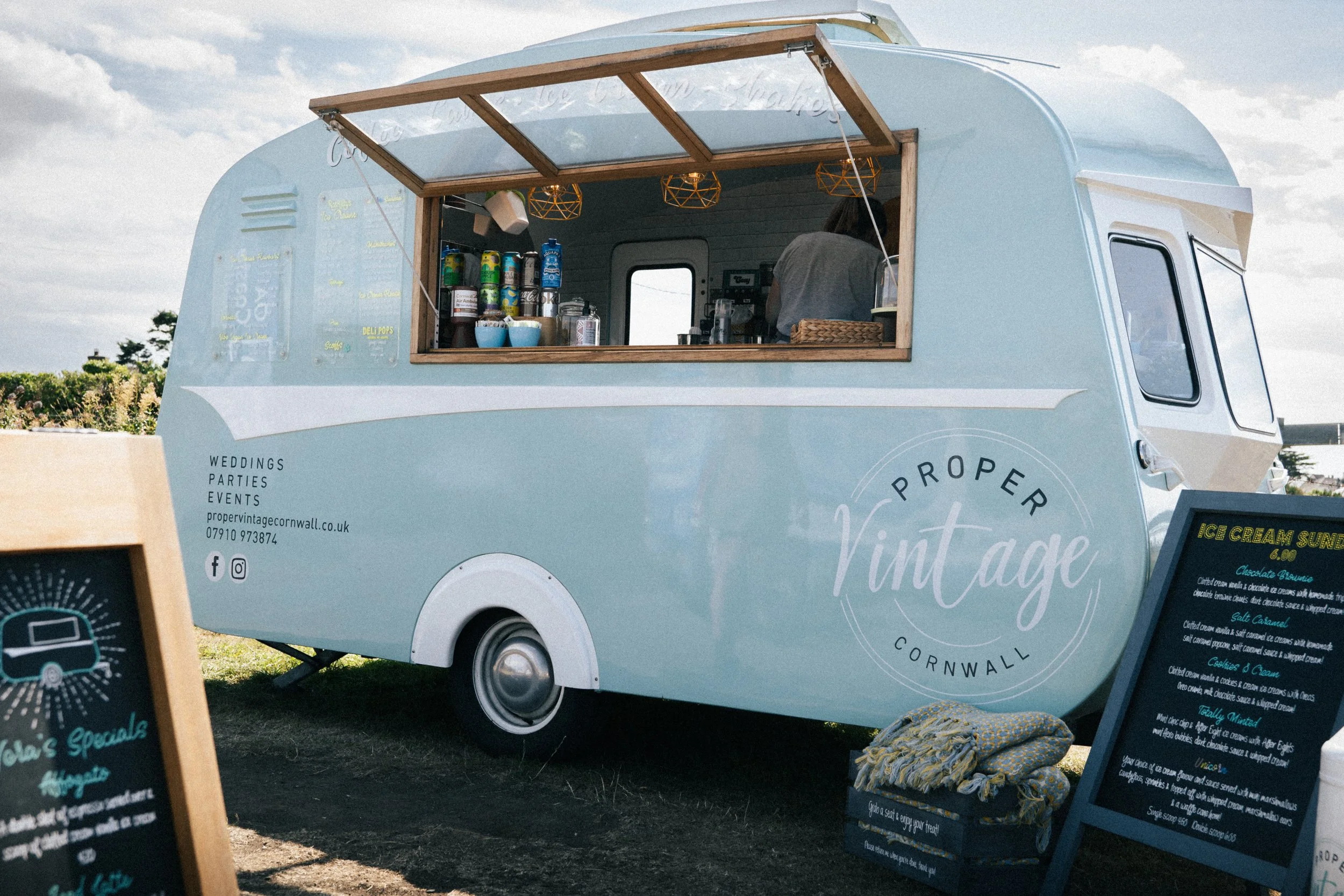 A light blue vintage mobile food trailer serving ice cream, with an open window showing a person inside preparing orders. Outside, there are chalkboard menus and promotional signs.
