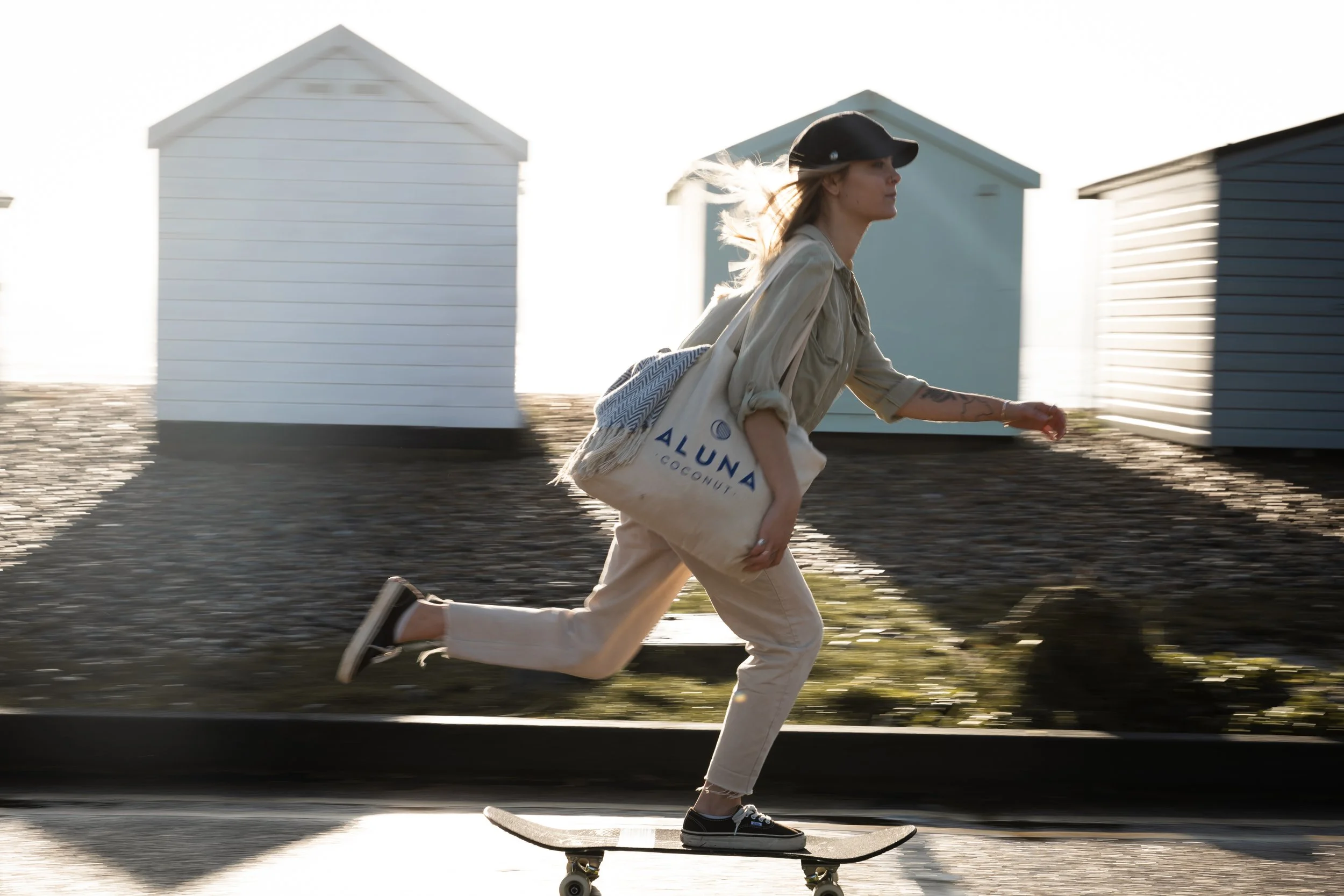 A woman skateboarding along the street with beach huts in the background, carrying a tote bag that says 'Aluna Coconut'.