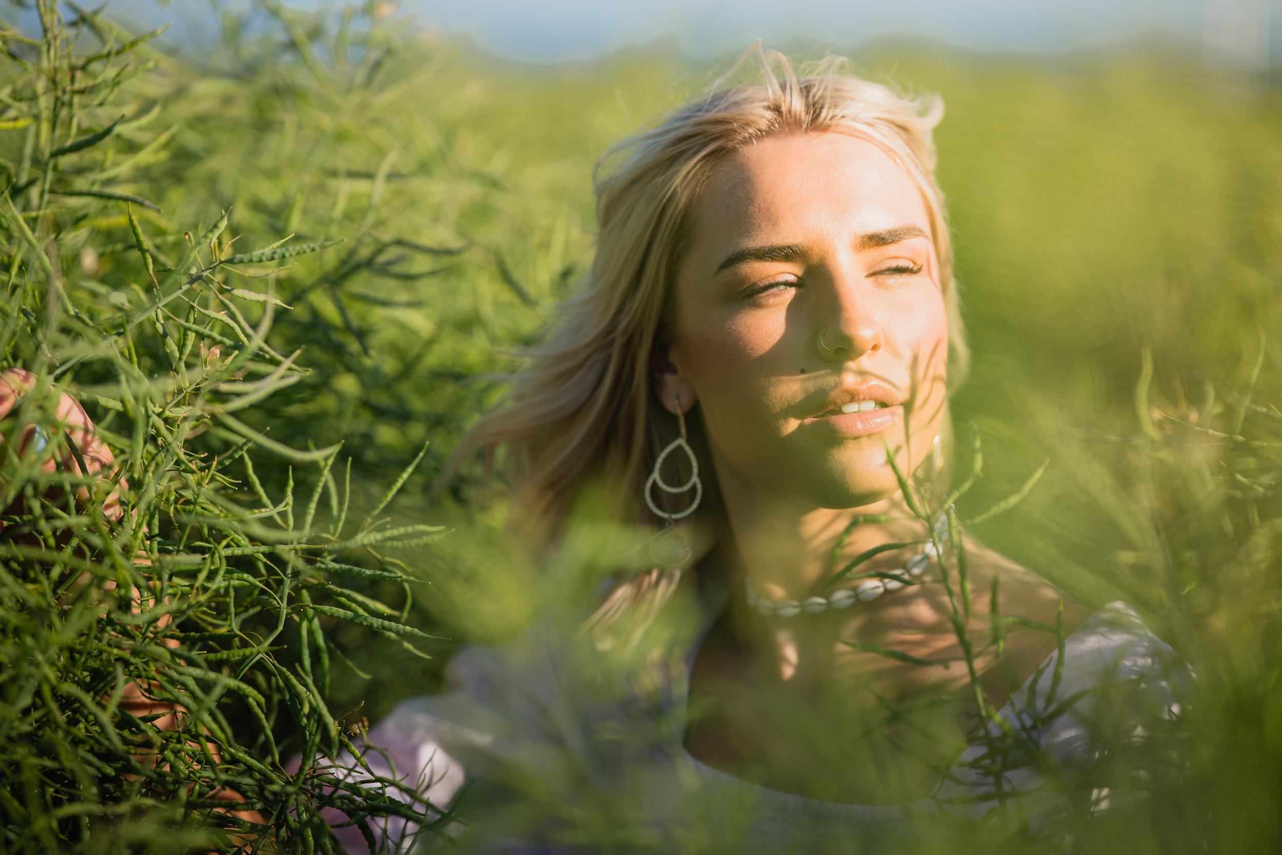 A woman with blonde hair and earrings with closed eyes sitting in a lush green field.