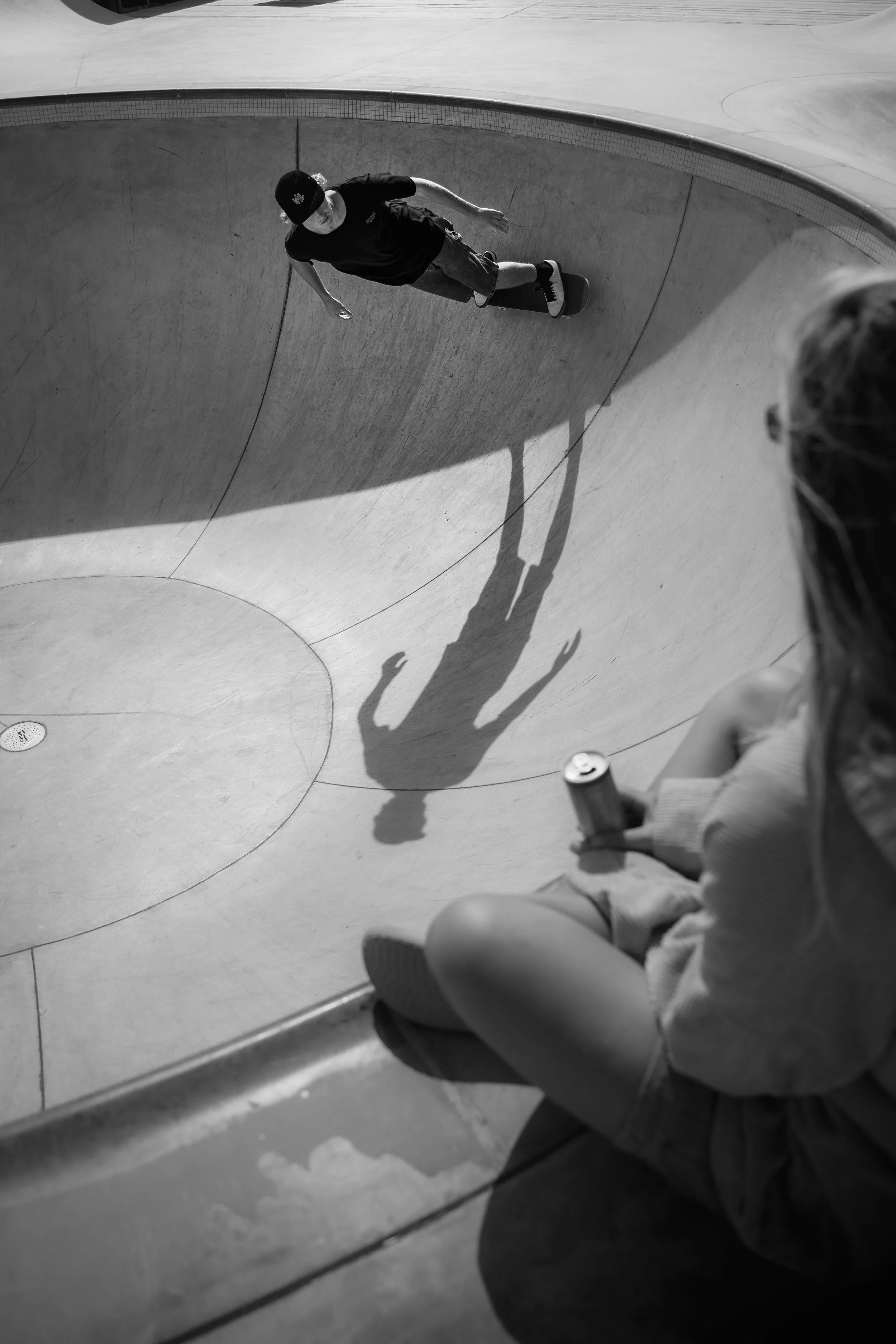 A person skateboarding in a skate park. The photo is taken from above, capturing the skateboarder’s shadow on the concrete and a girl sitting on the edge of the skate bowl holding a can.