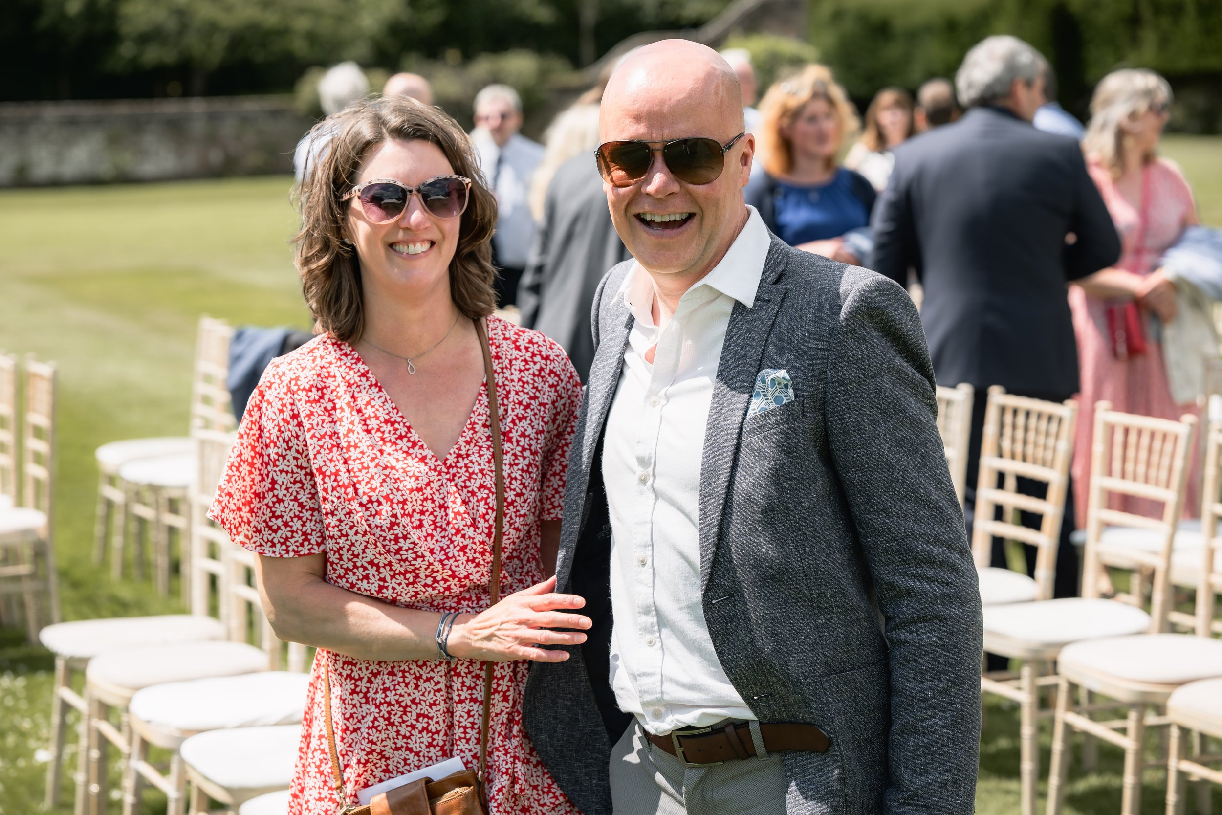 A smiling woman in a red patterned dress and sunglasses standing next to a smiling man in a gray blazer and sunglasses at an outdoor event with chairs and other guests in the background.