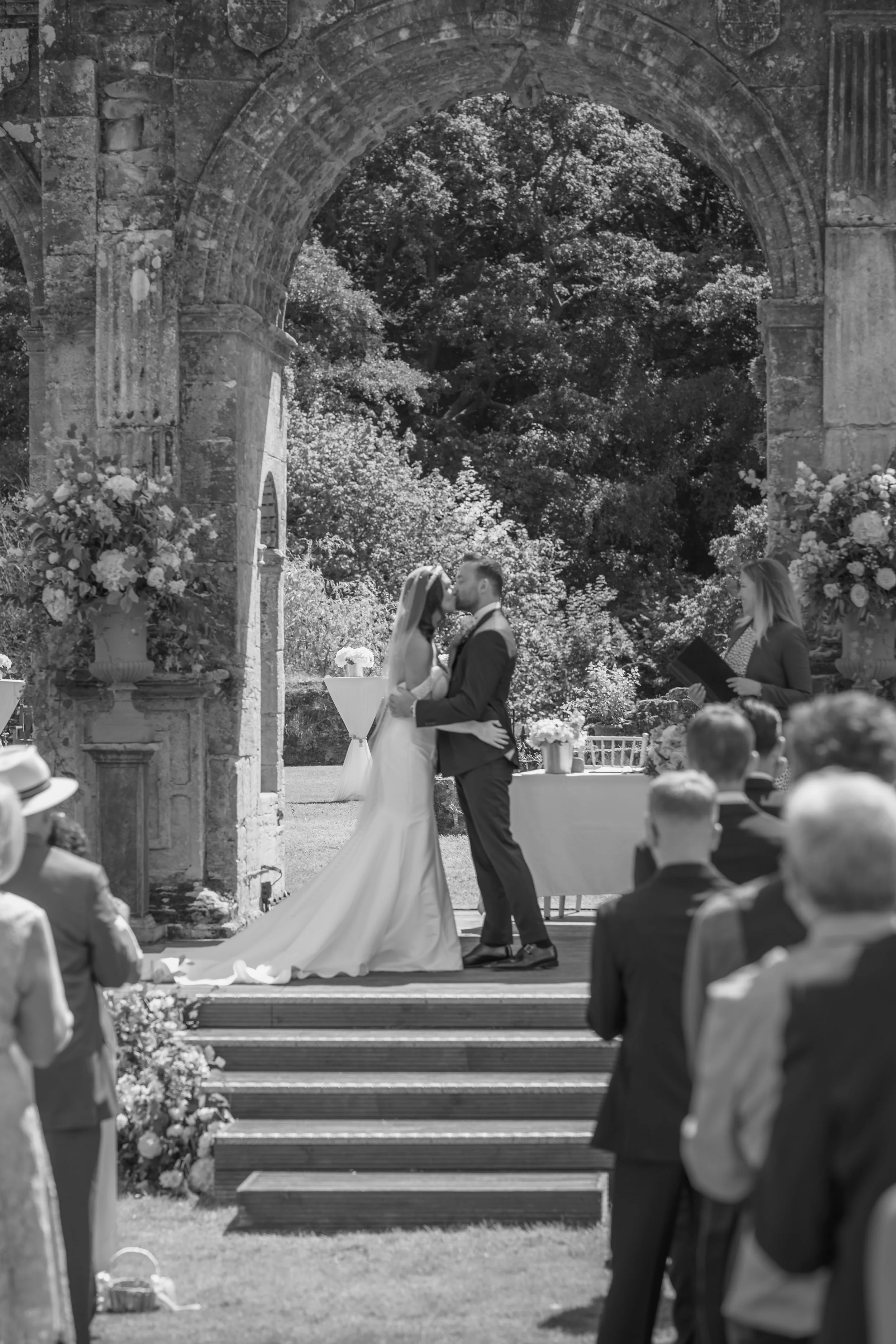 A black and white photo of a wedding ceremony outdoors with a bride and groom kissing under an arch with guests watching.