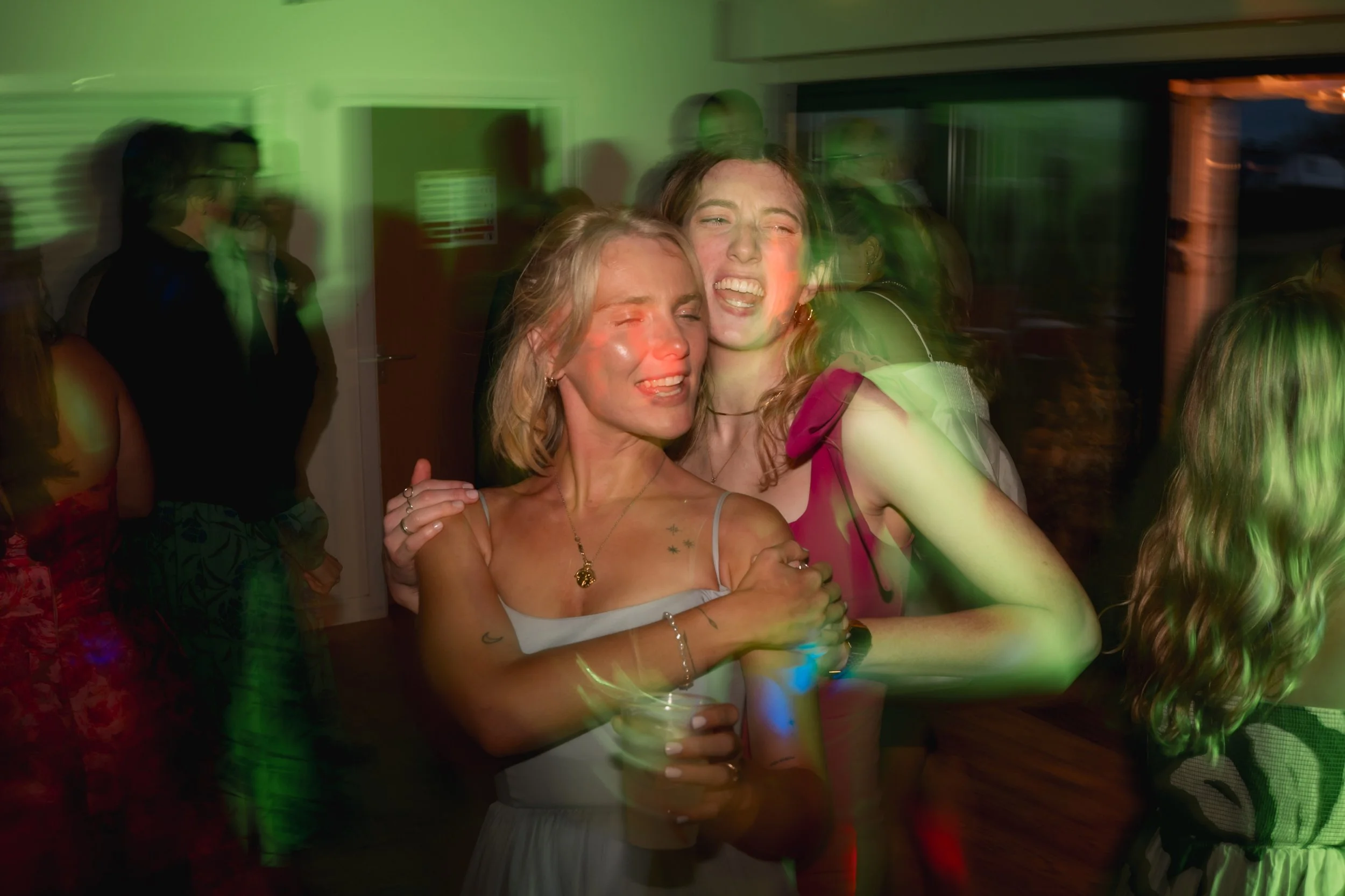Two women hugging and smiling at a lively party or gathering, with colorful lights and other people visible in the background.