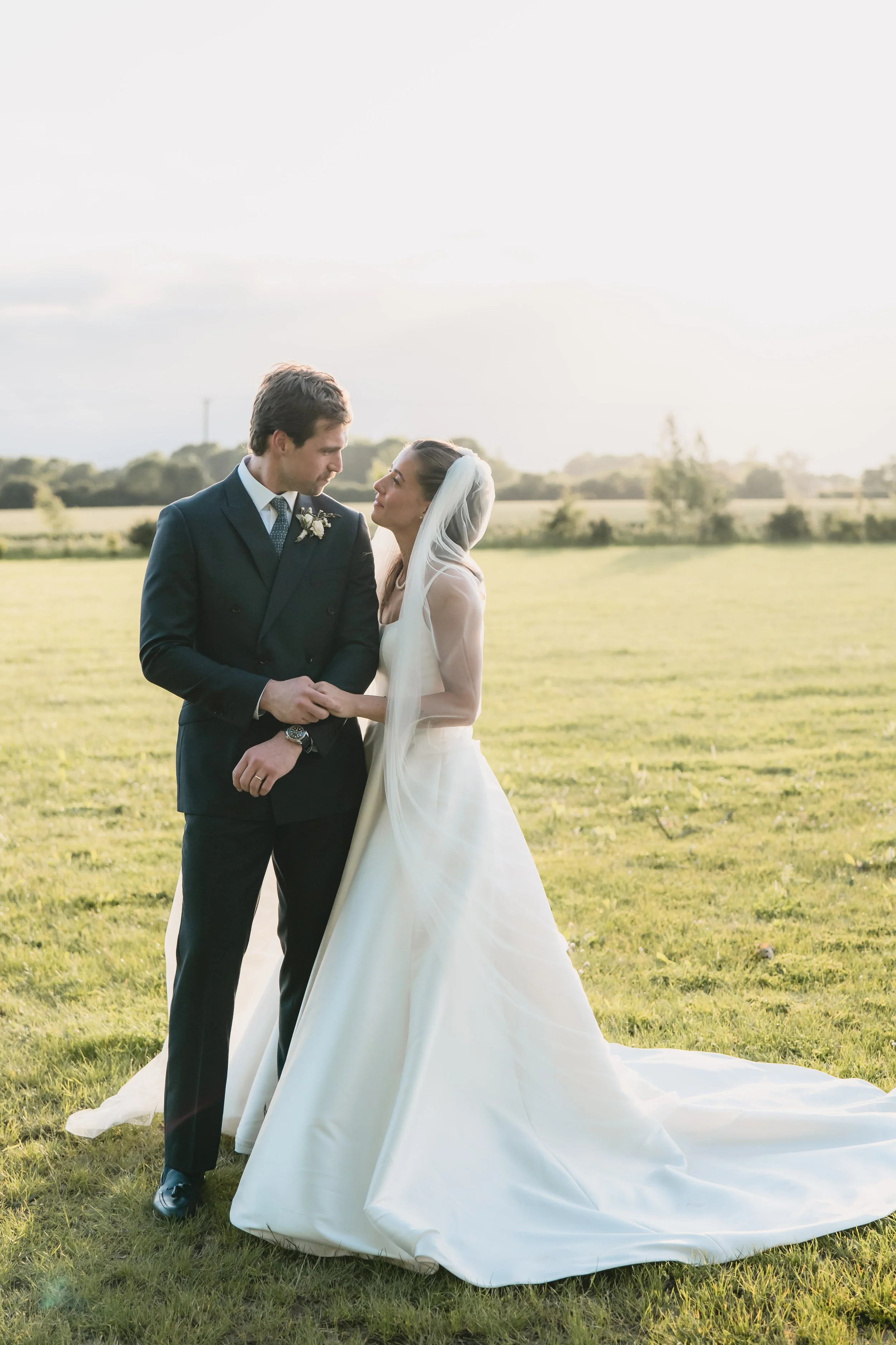 A bride and groom standing together in a field during sunset, holding hands and gazing at each other.