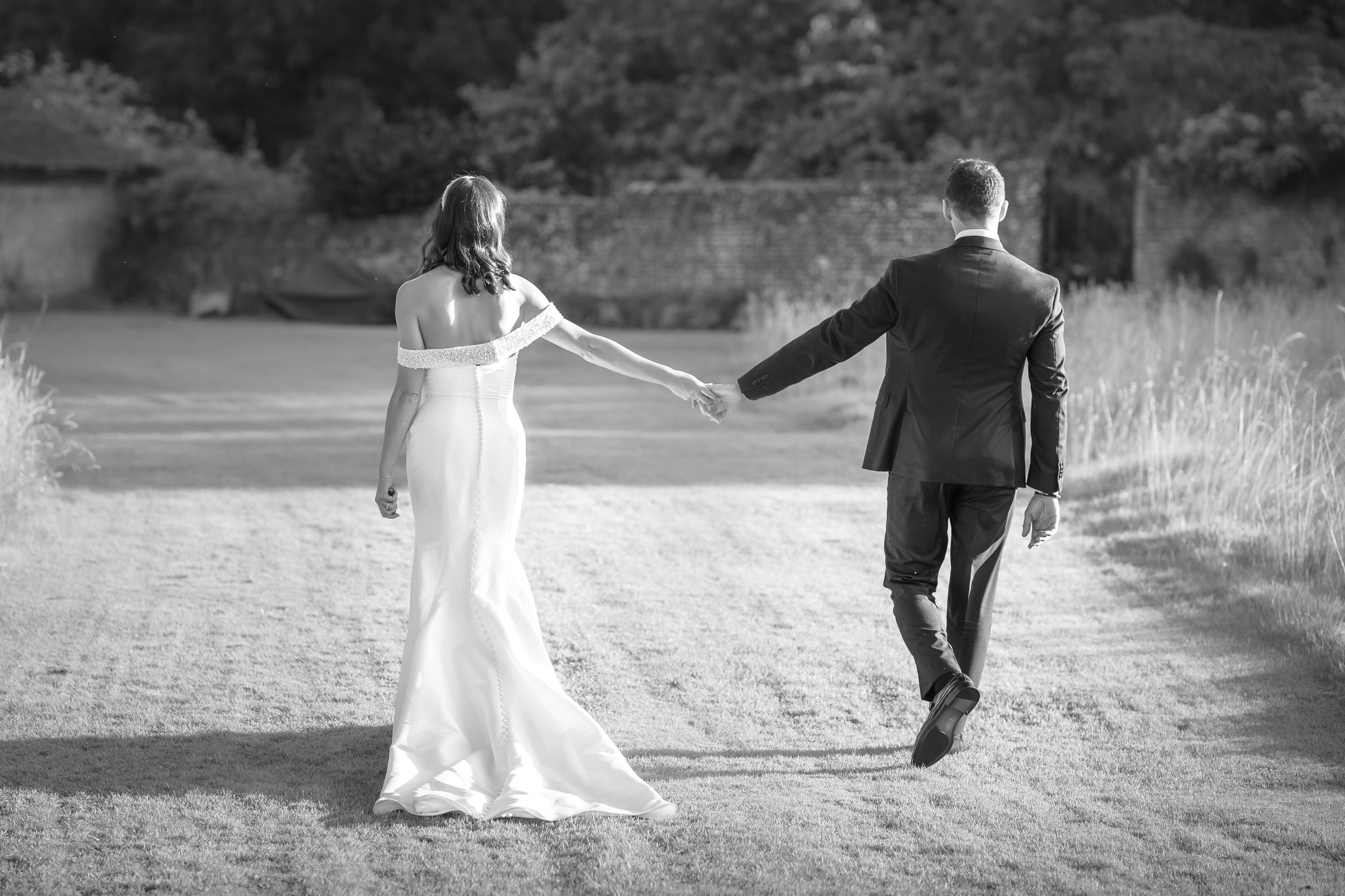 A bride and groom holding hands and walking away outdoors, wedding attire, natural background, black and white photograph.