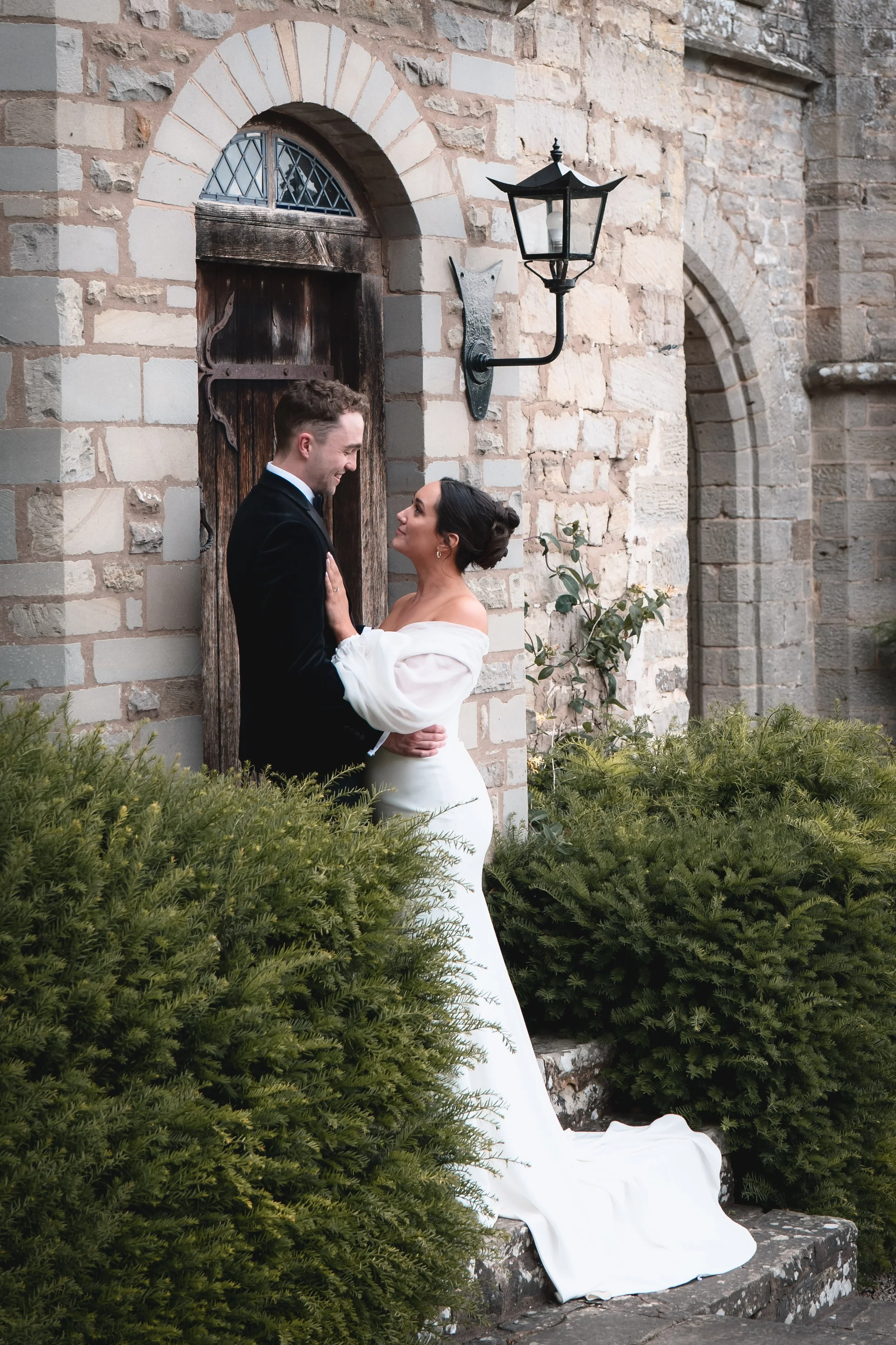A bride and groom standing close together outside a stone building, smiling at each other, surrounded by greenery.