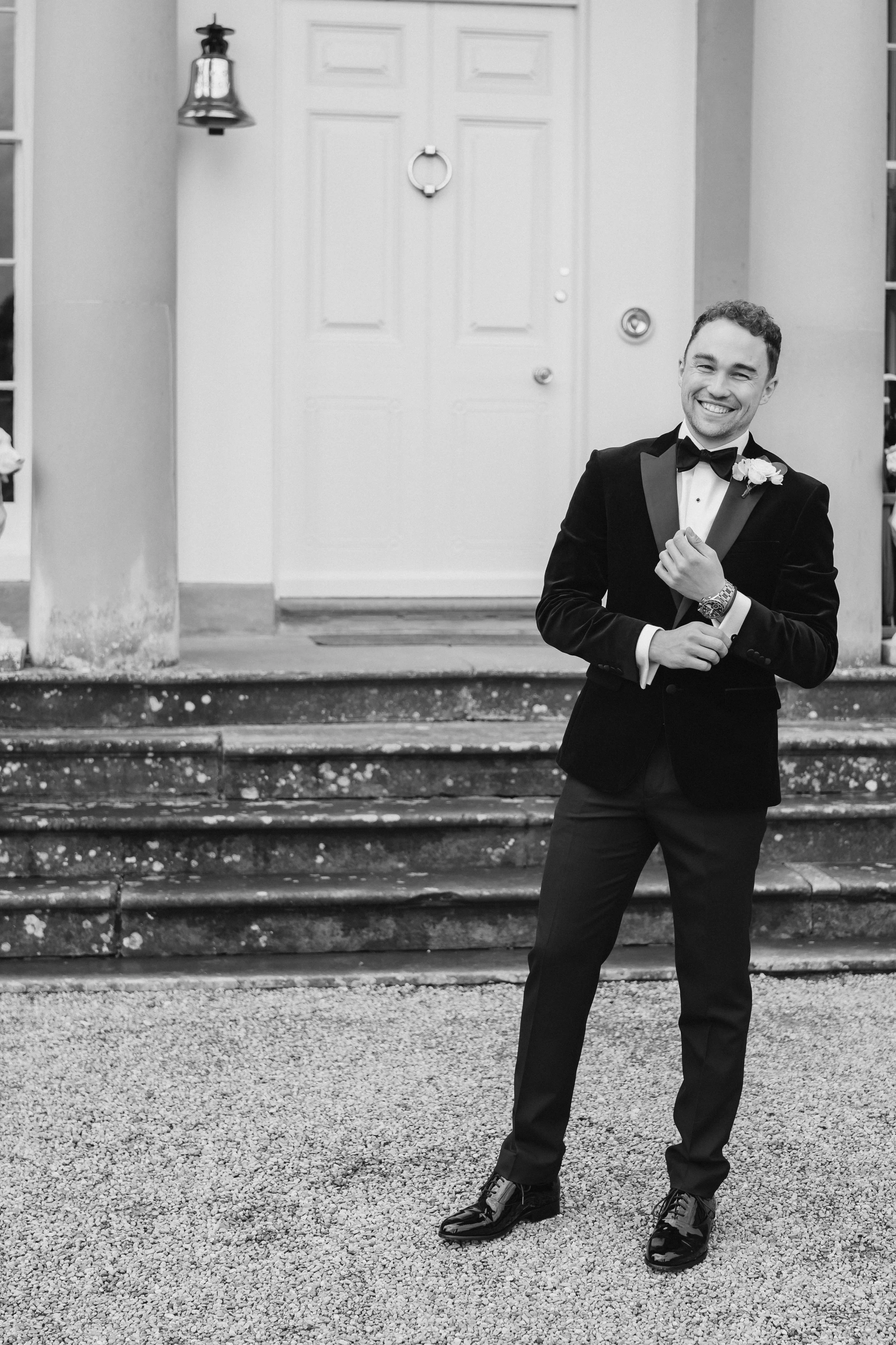 A man in a tuxedo with a bow tie, smiling and standing outside a house with steps, a door, and a lantern on the wall.