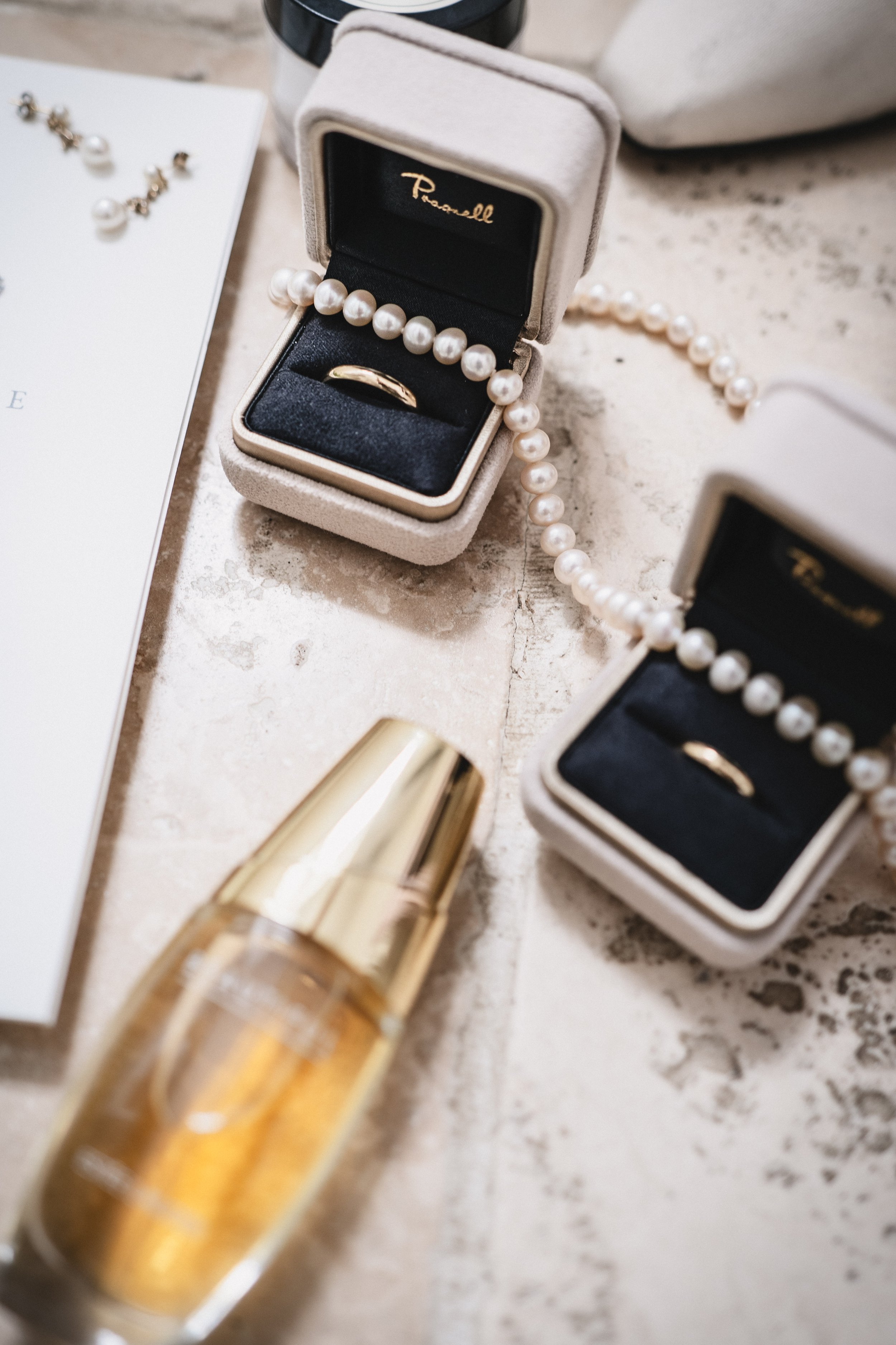 Two jewelry boxes with pearl necklaces and a gold ring inside, a gold perfume bottle, and part of a white paper or card on a textured surface.