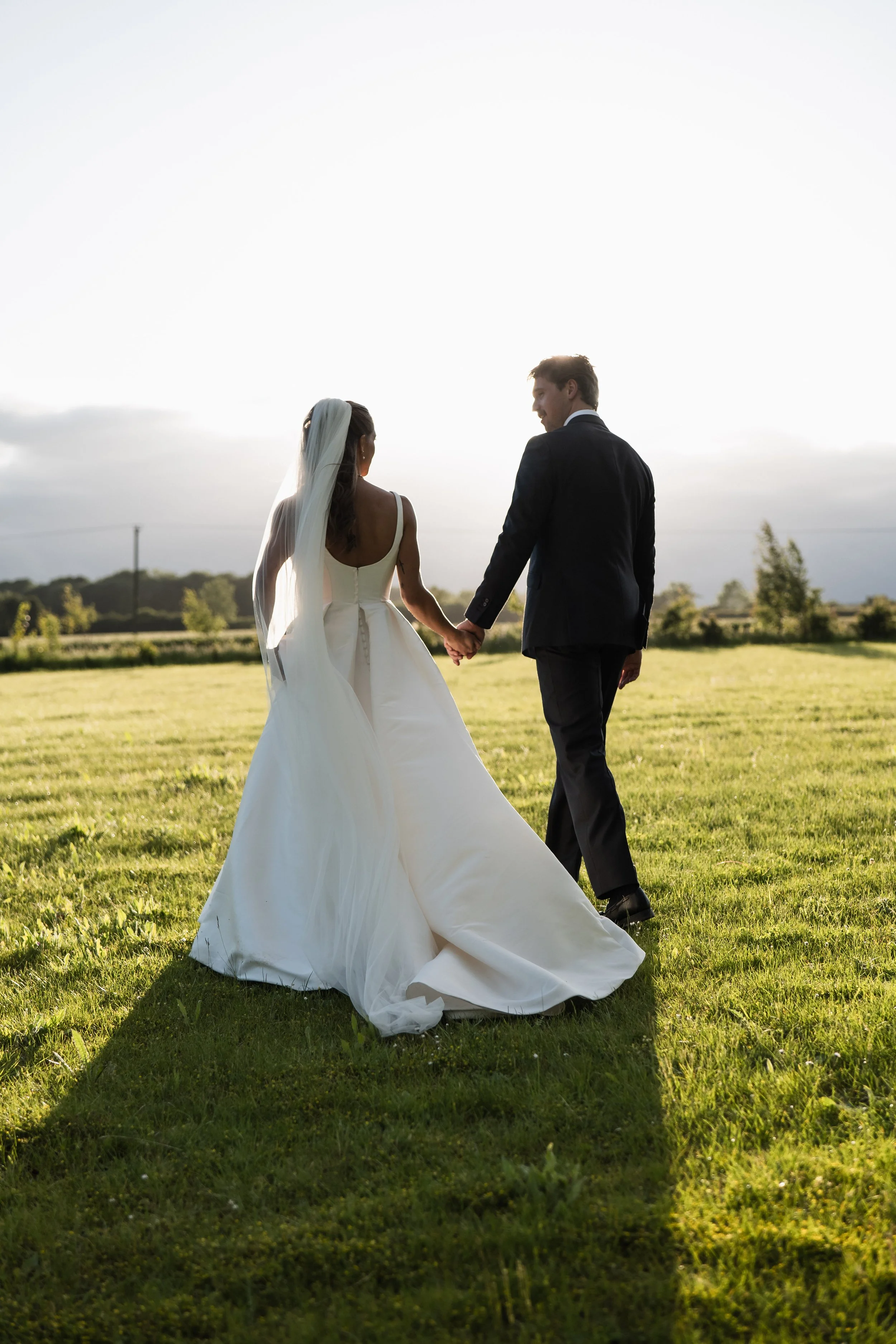 A bride and groom holding hands and walking on a grassy field during sunset, facing away from the camera.