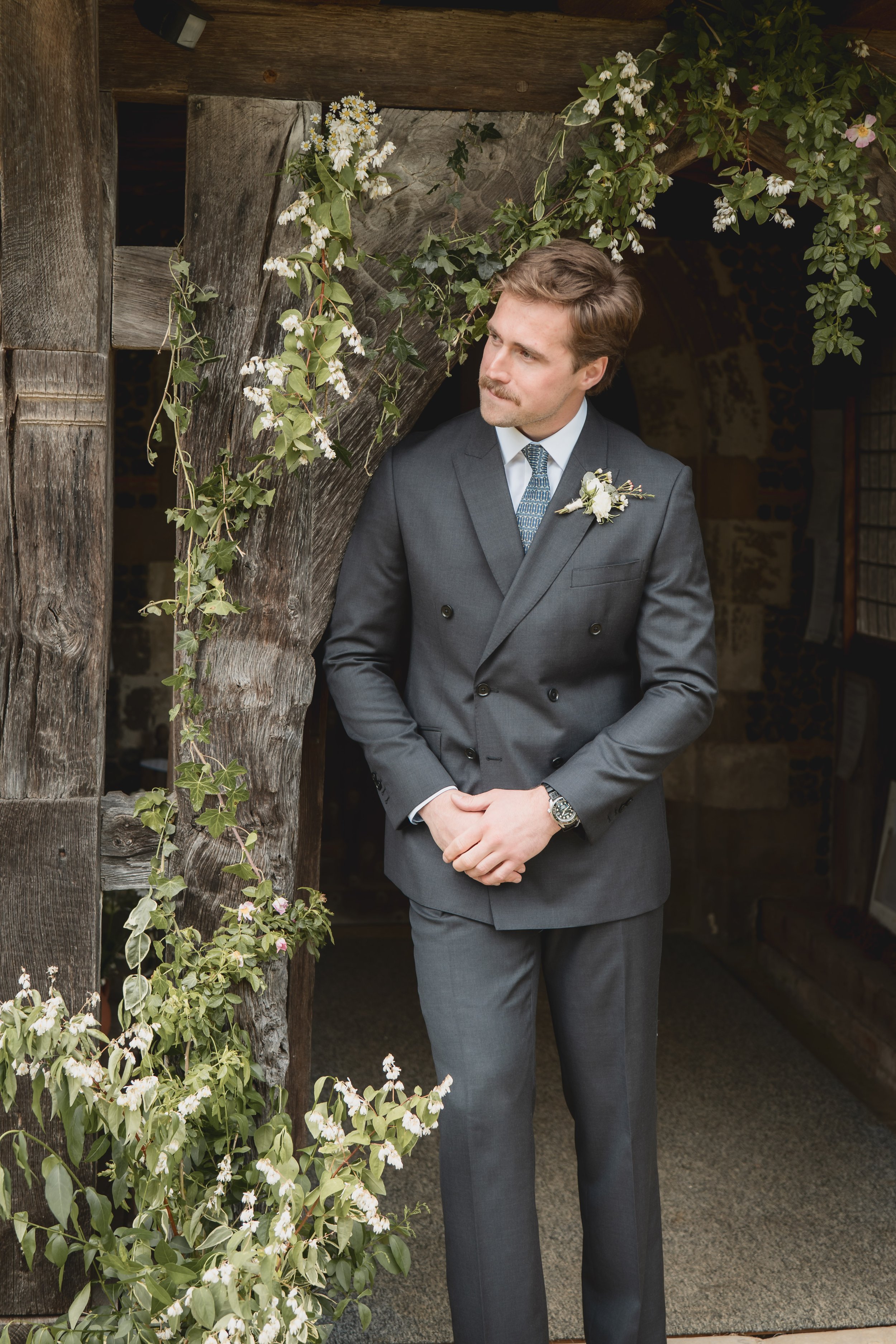 A man in a gray suit stands at a rustic wooden doorway decorated with green ivy and white flowers, looking to the side with a contemplative expression.