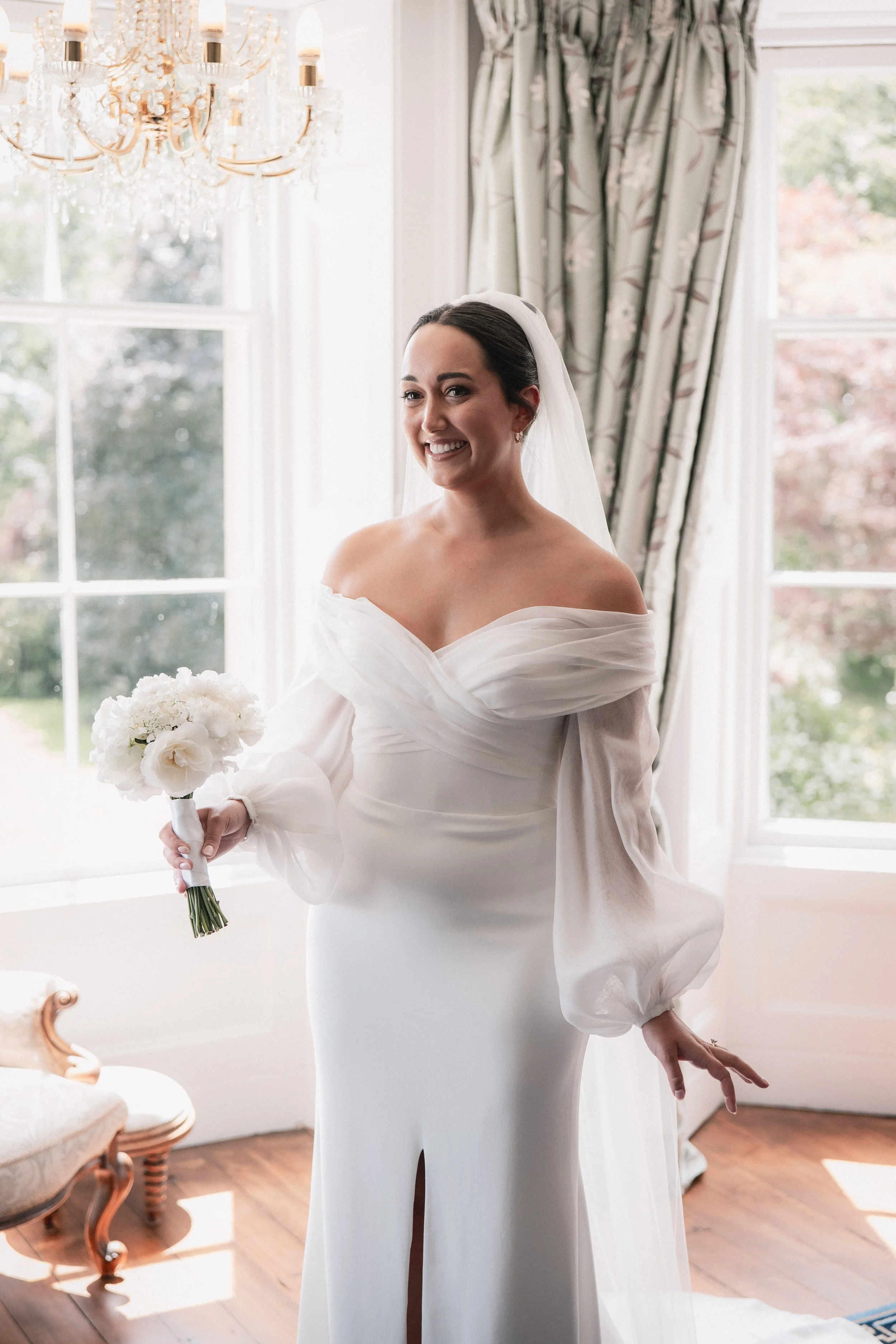 A bride with dark hair in a white off-shoulder wedding dress holding a small bouquet of white flowers, standing in a bright room with large windows and floral curtains, smiling.