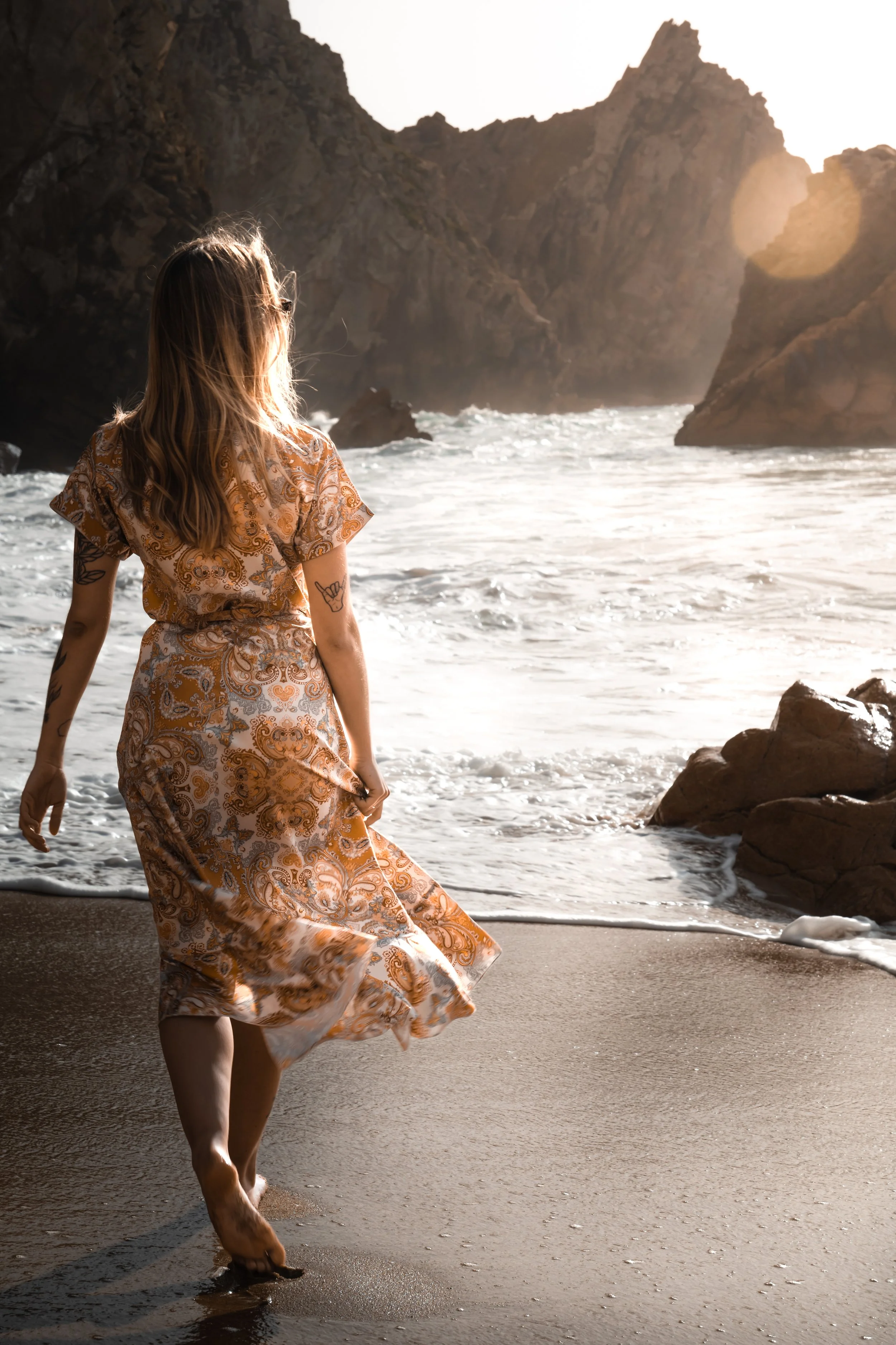 A woman with tattoos, wearing a flowing, patterned dress, walking barefoot on a sandy beach at sunset with rocky cliffs in the background.
