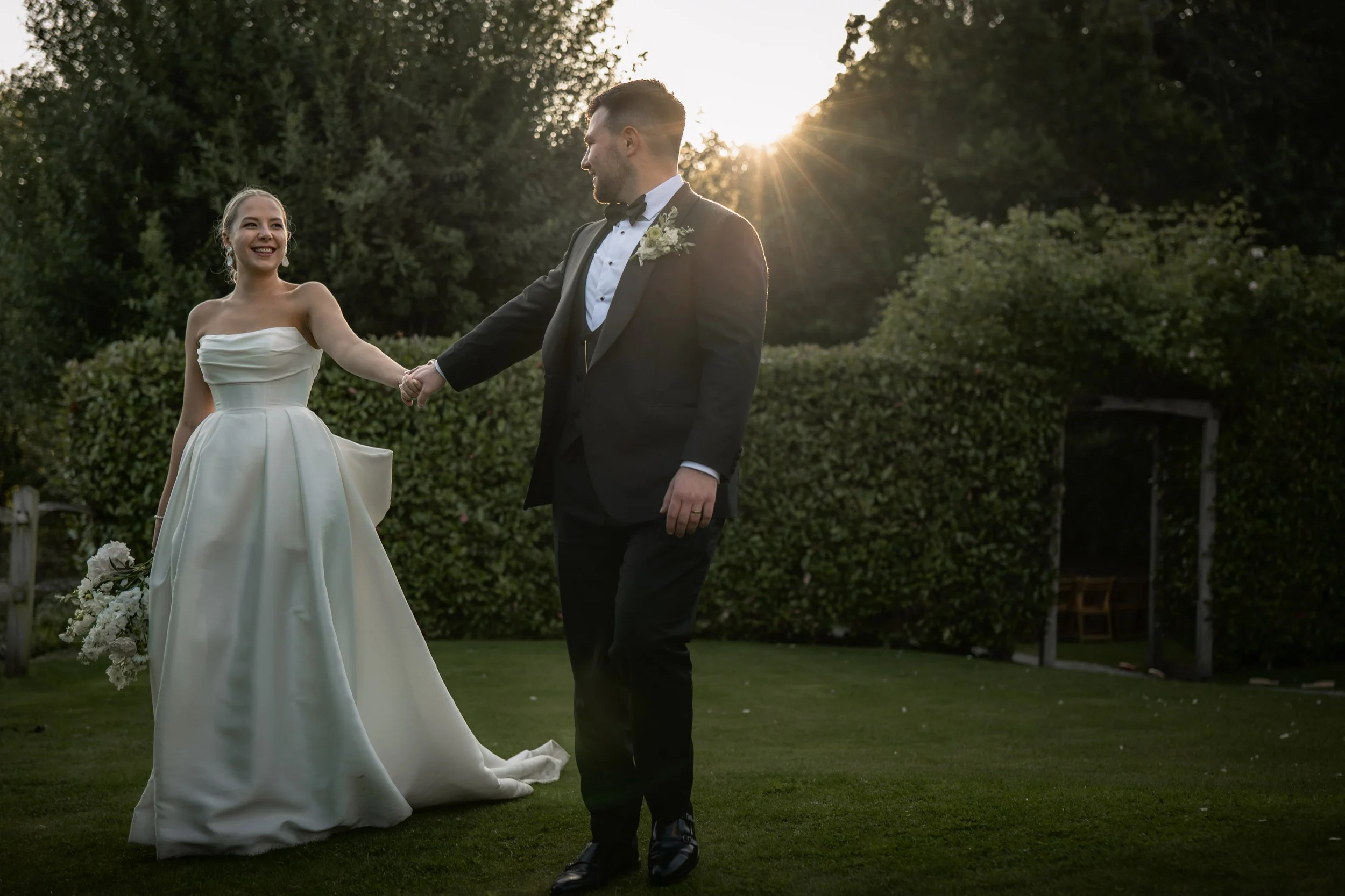 A bride and groom holding hands outdoors during sunset. The bride is in a strapless white wedding gown holding a bouquet, smiling at the groom. The groom is dressed in a black tuxedo with a bow tie, holding the bride's hand. Sun rays shine behind the