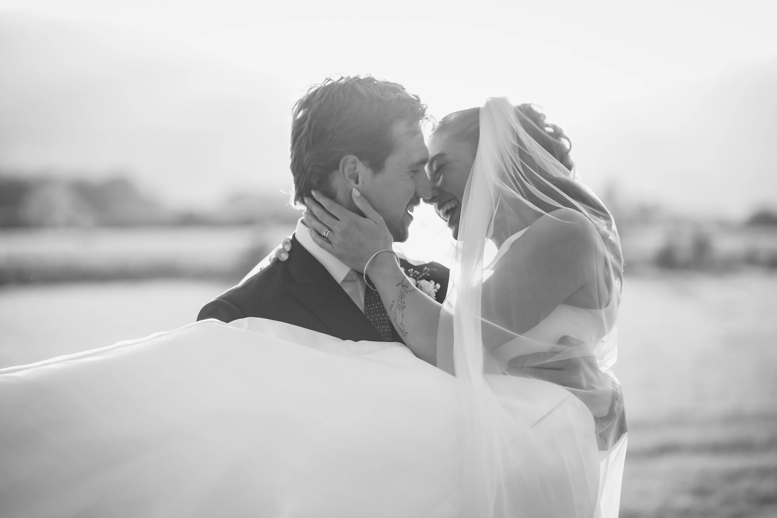 A black-and-white photo of a bride and groom smiling and touching foreheads, outdoors, with the bride wearing a veil and the groom in a suit.