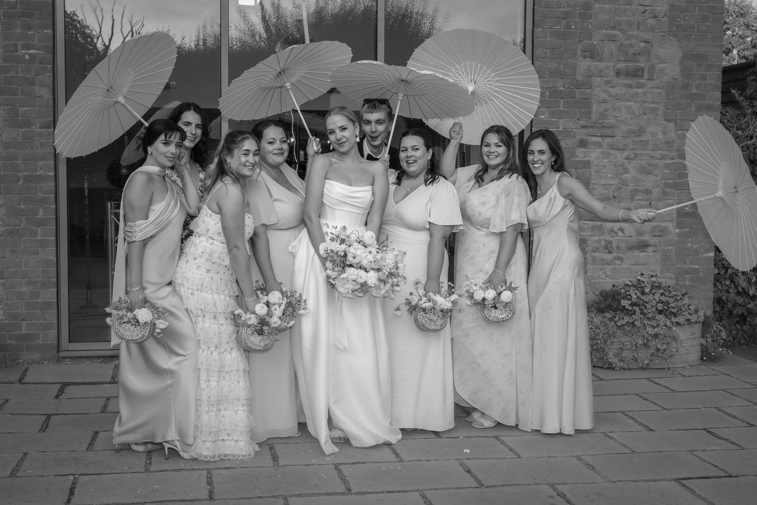 Black and white photograph of a wedding party standing outdoors in front of a brick building with large windows. The group includes the bride holding a bouquet of flowers, surrounded by several bridesmaids holding parasols and bouquets. The women are