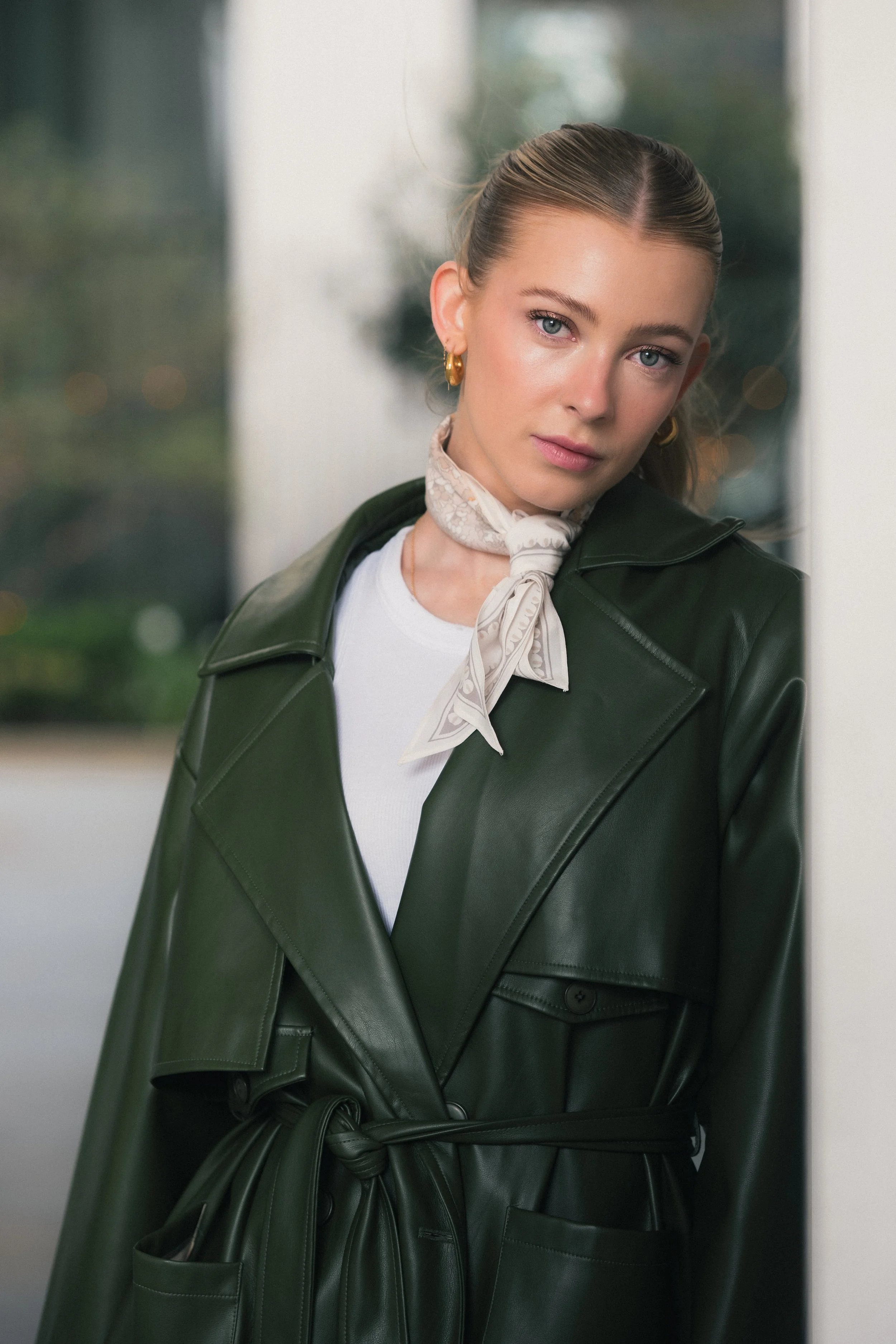 A woman with blonde hair, blue eyes, wearing gold hoop earrings, a white shirt, a beige scarf around her neck, and a green leather trench coat, standing by a window with a blurred outdoor background.