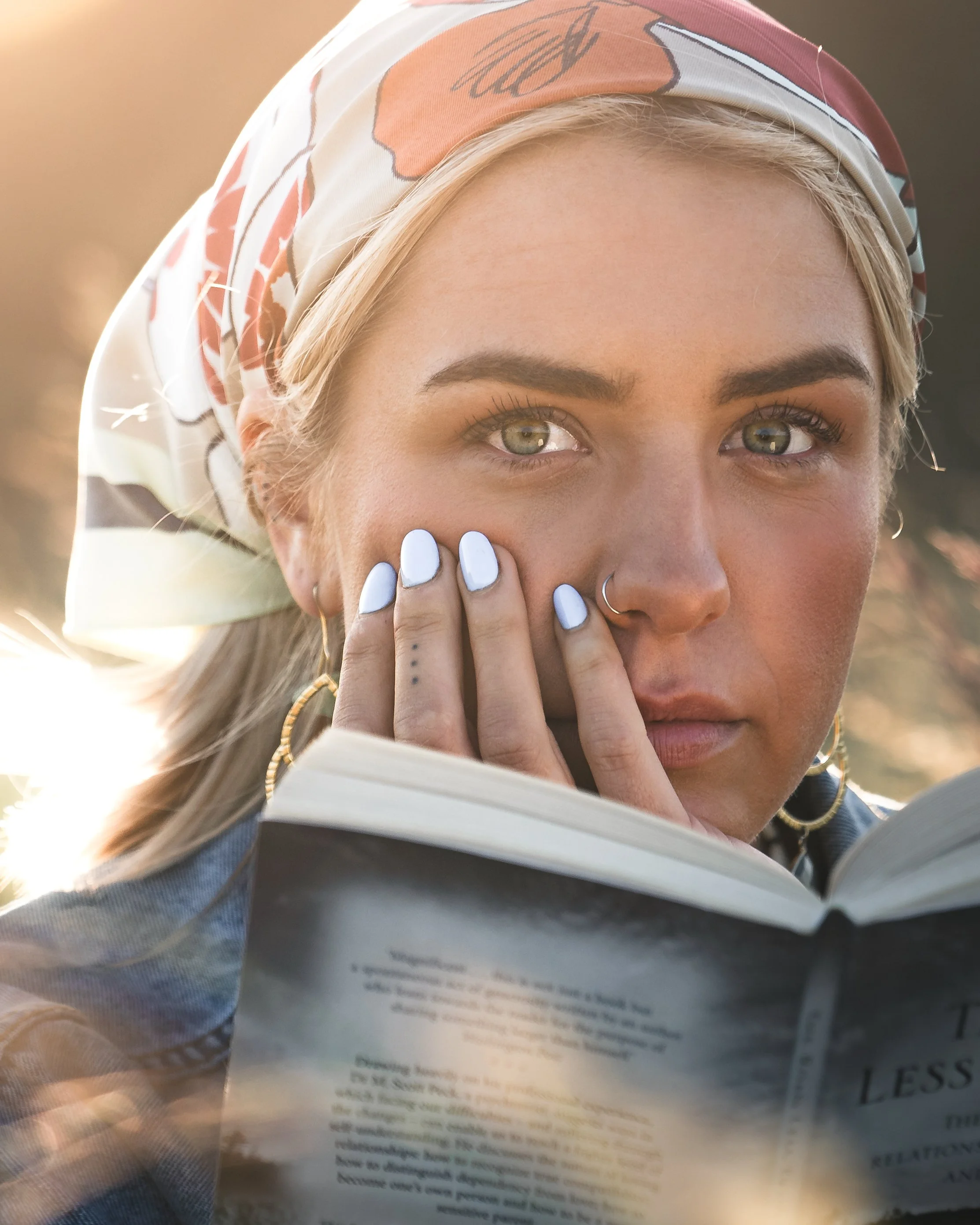 Close-up of a woman with blonde hair, wearing a patterned headscarf, holding a book, with her fingers near her face, wearing white nail polish and a nose ring, outdoors during sunset.