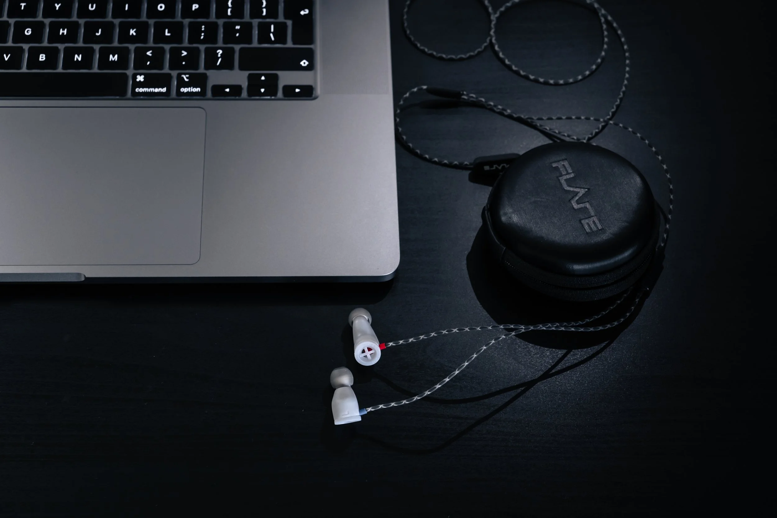 Close-up of a laptop keyboard and touchpad, with a pair of in-ear headphones and a small round headphone case on a dark surface.