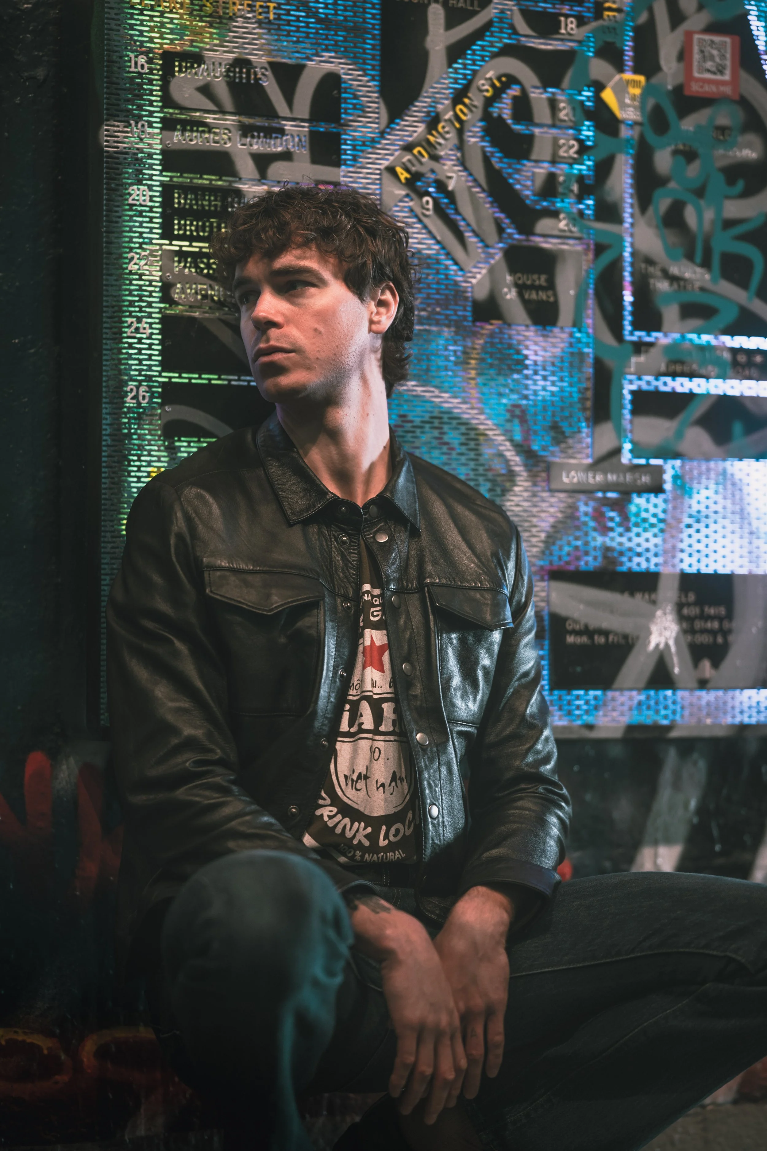 A young man with curly hair wearing a black leather jacket and graphic t-shirt sitting against a colorful, graffiti-covered wall with neon lights.