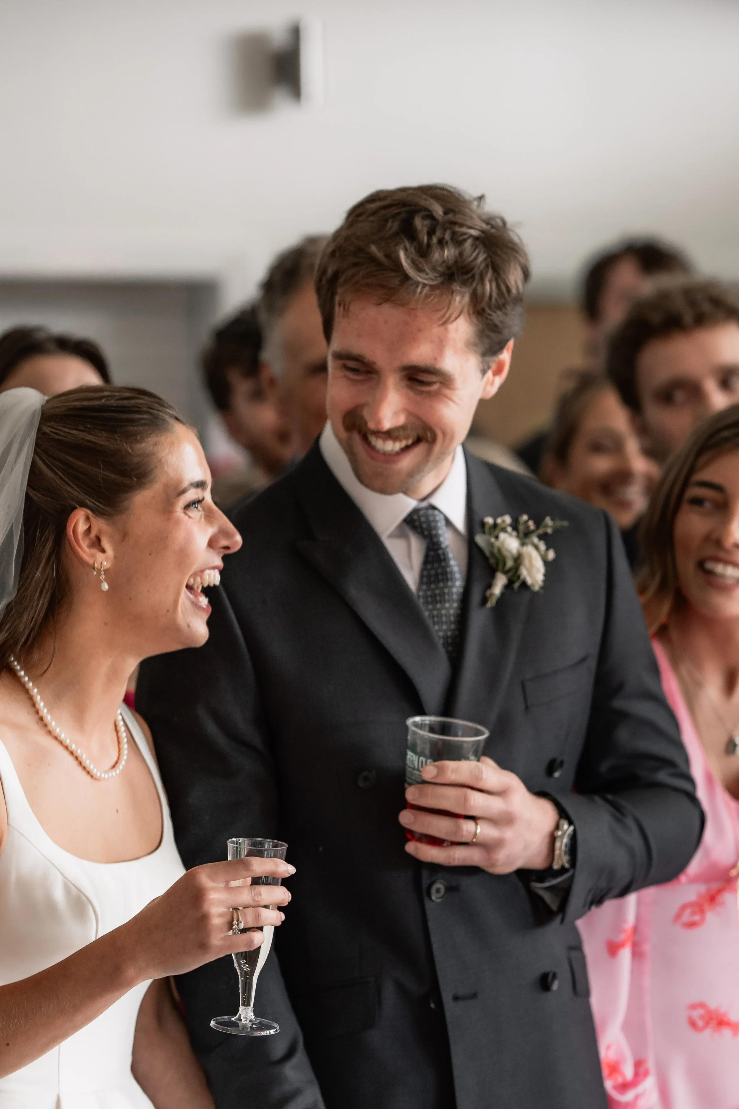 A woman in a white dress and pearls is smiling and holding a glass of champagne while talking to a man in a suit with a boutonnière, also holding a glass. They are at a wedding celebration with other guests in the background.