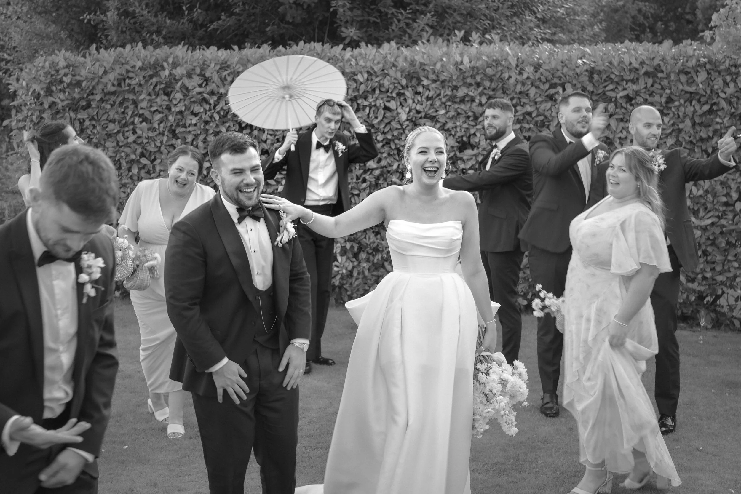 A black-and-white photo of a wedding celebration outdoors, featuring the bride and groom laughing and surrounded by friends and family.