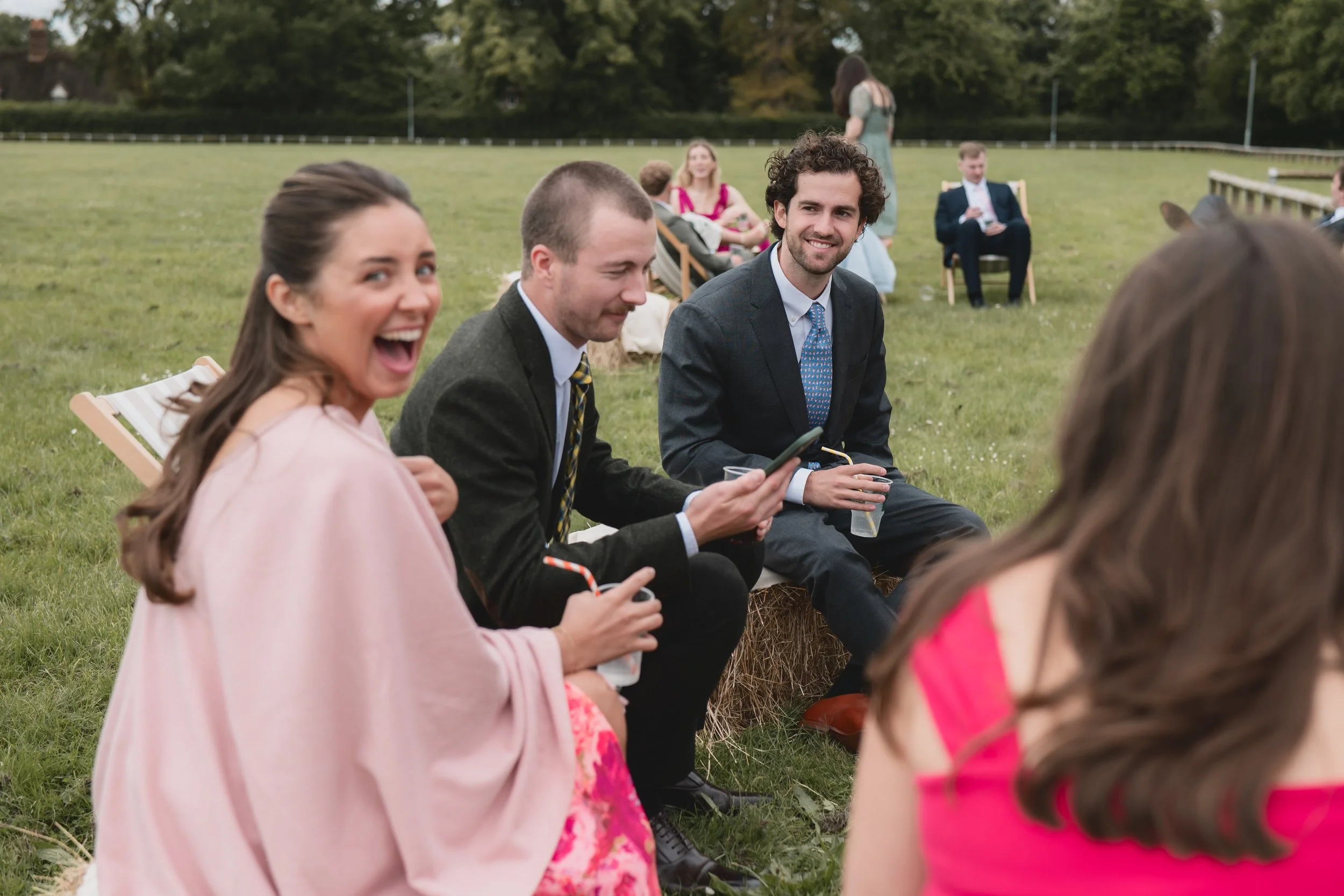 Group of people sitting on hay bales at an outdoor event, laughing and enjoying drinks, with a grassy field and trees in the background.