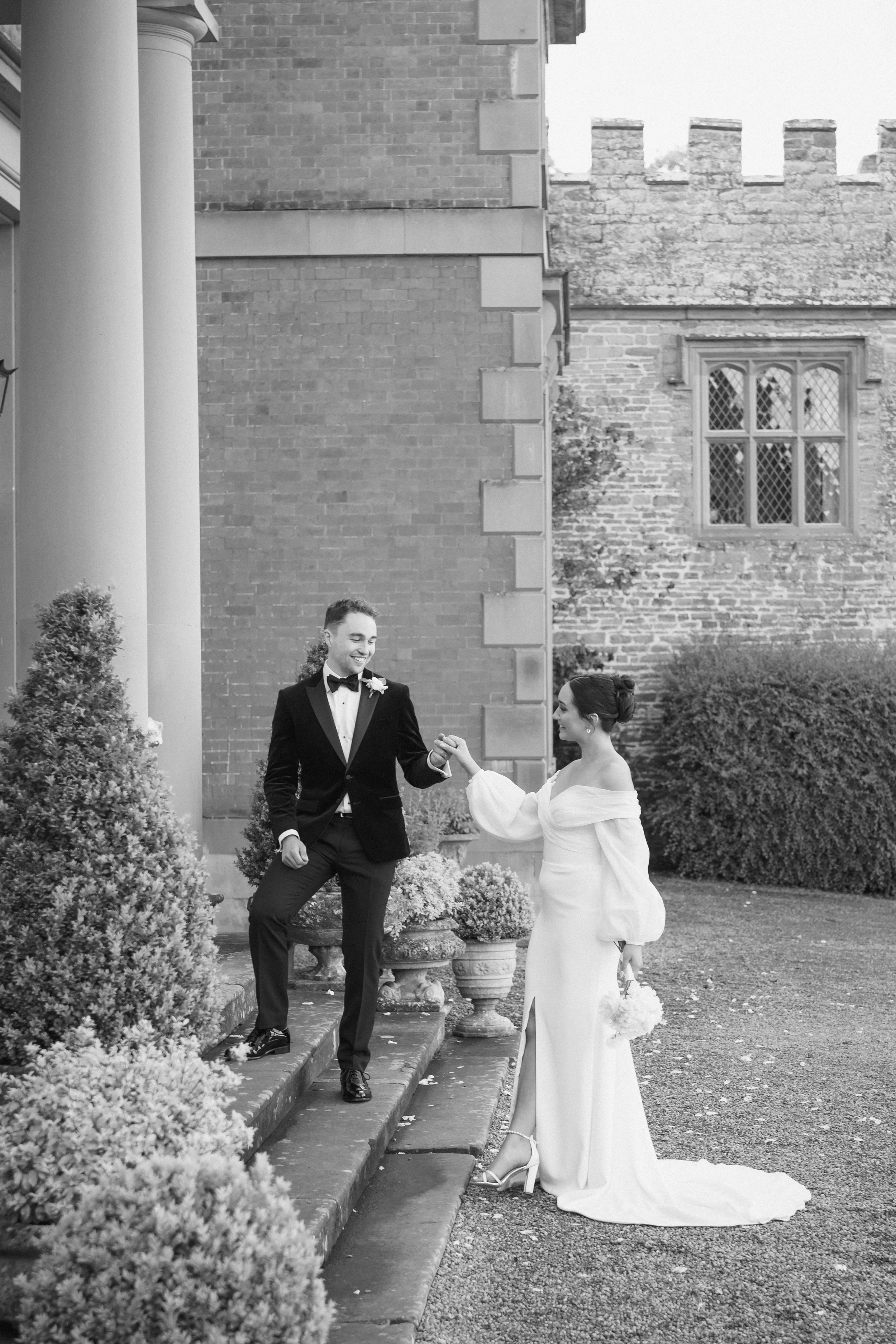 A black and white photo of a bride and groom outdoors; the groom is on stairs holding the bride's hand, both smiling, near a brick building with tall windows and greenery.