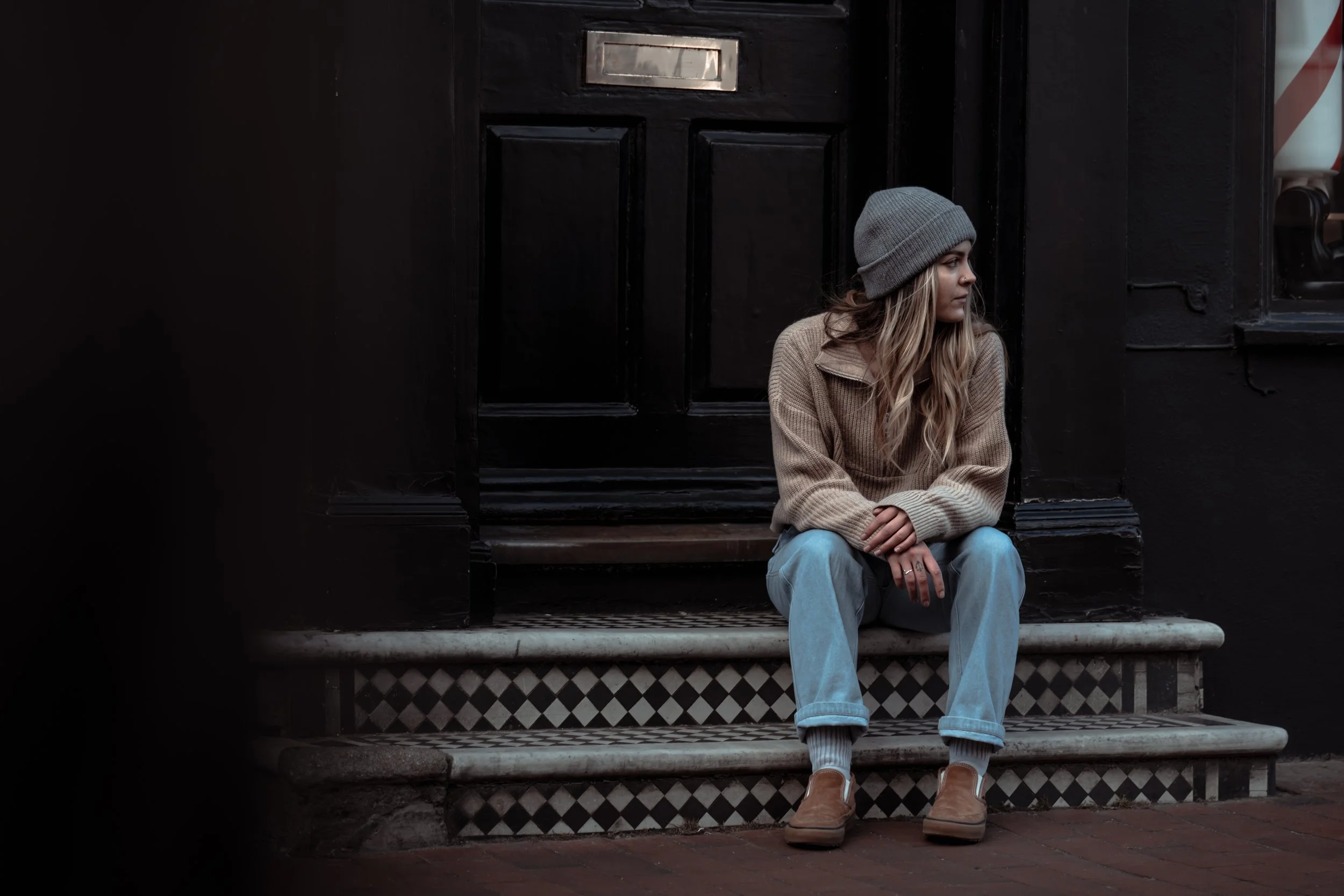A young woman with long blond hair sitting on the steps in front of a black door, wearing a gray beanie, beige sweater, light blue jeans, and tan shoes, looking to the side.