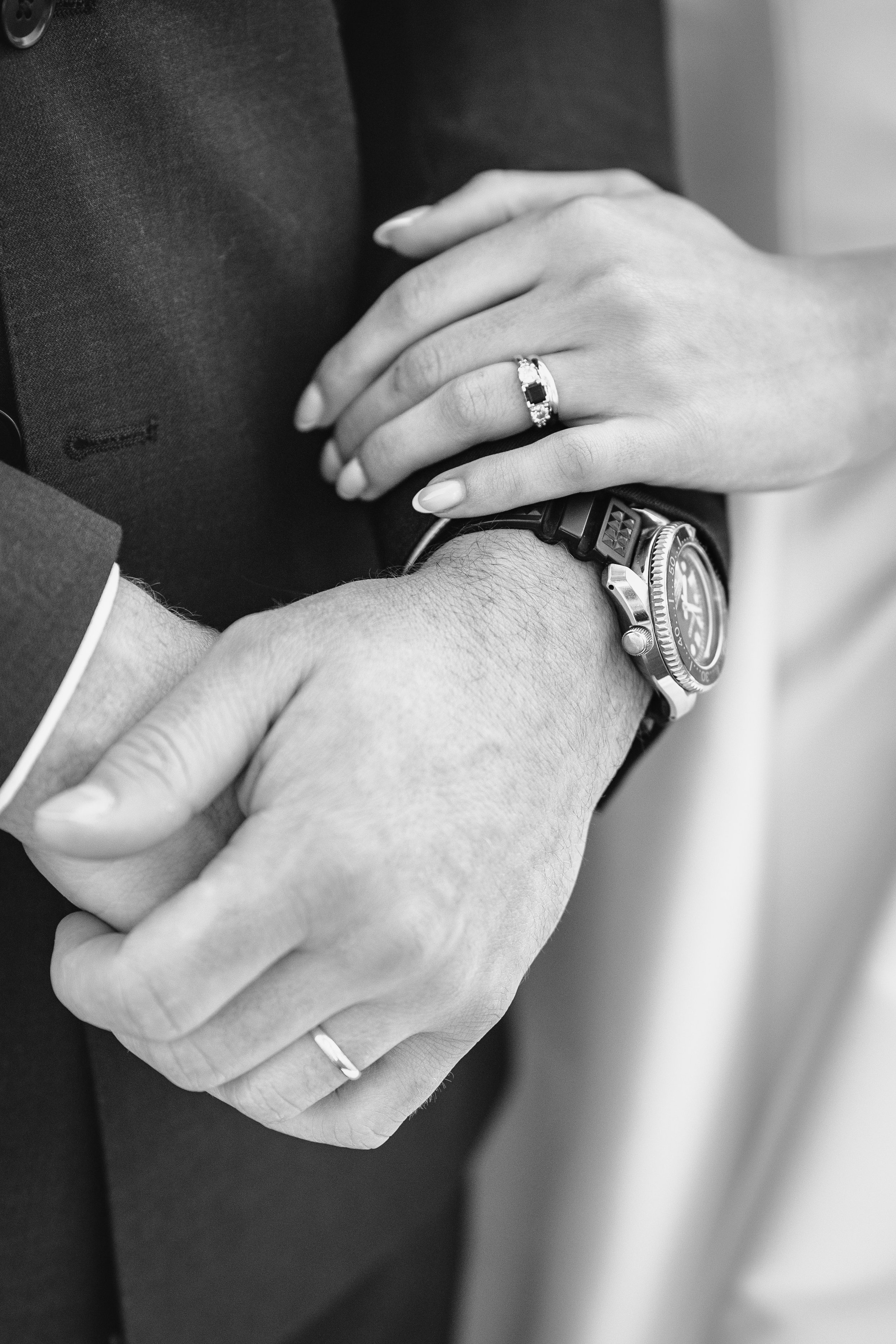 Close-up of a couple's hands intertwined, with the woman's hand resting on a man's wrist, showcasing wedding and engagement rings. The man wears a watch.