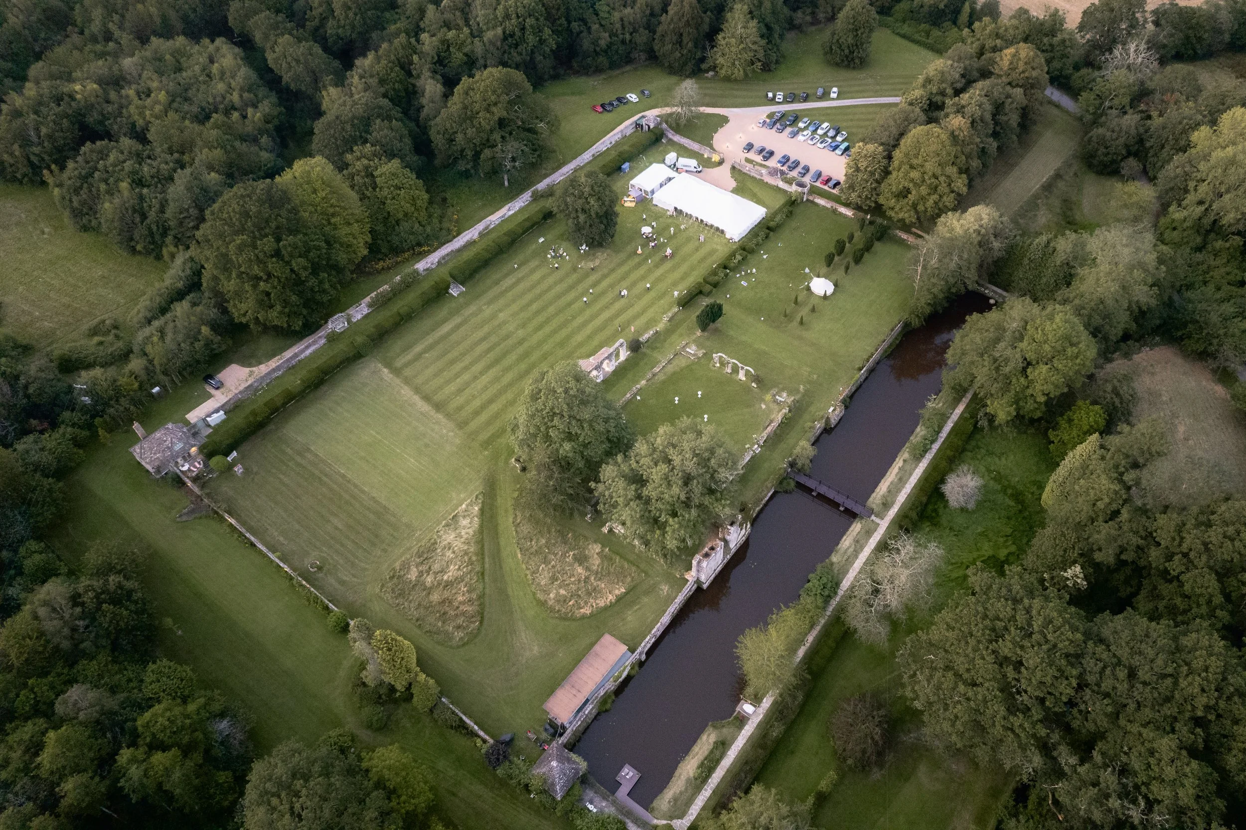 Aerial view of a large estate with manicured lawns, trees, a river running through it, a bridge over the river, and a parking lot. There is a large white event tent and several smaller structures, with people visible on the grounds.