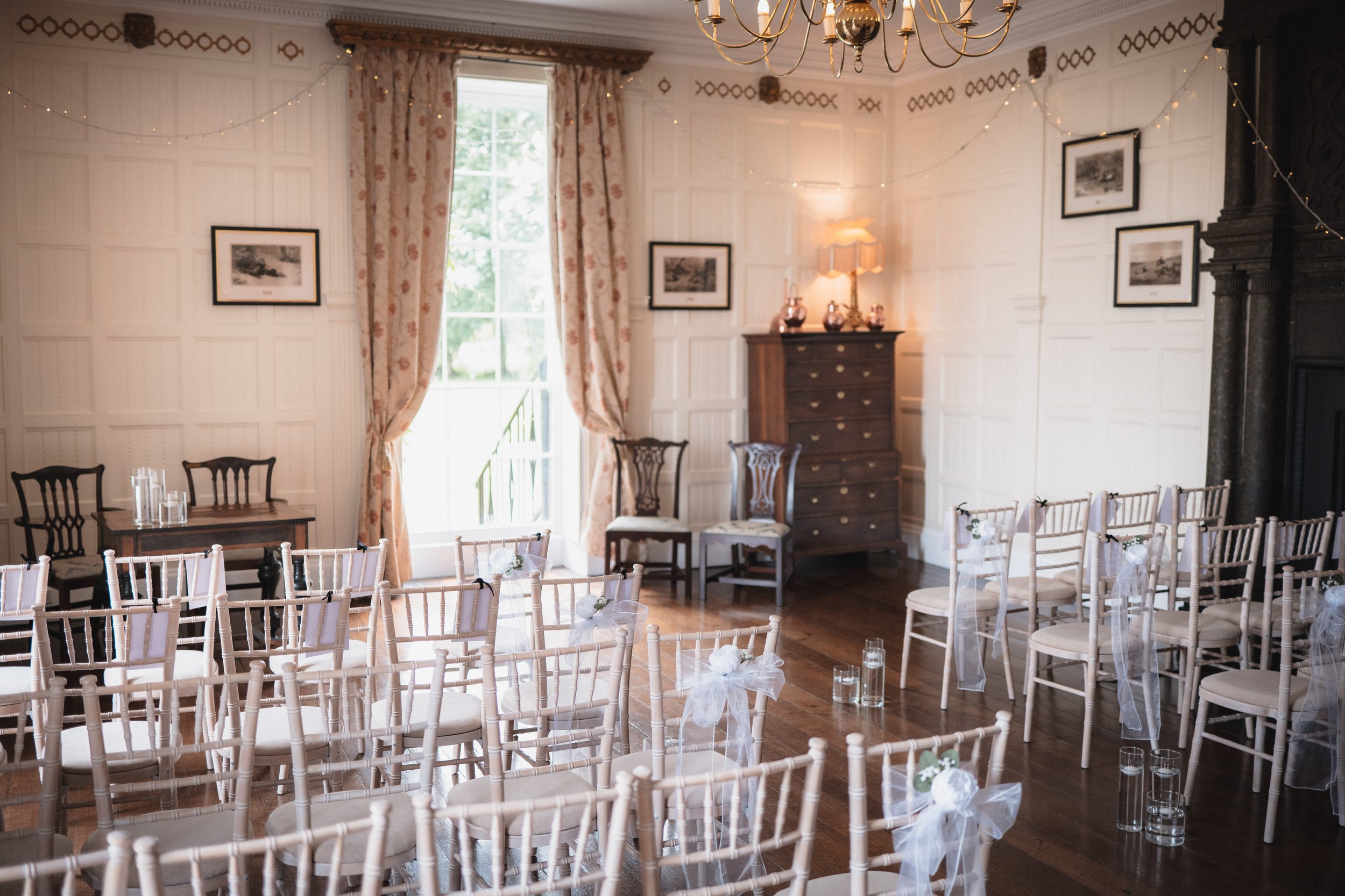 Indoor wedding ceremony setup with cream chairs decorated with white bows, small tables with glasses of water, and a large window with floral curtains in a decorated, classic room.