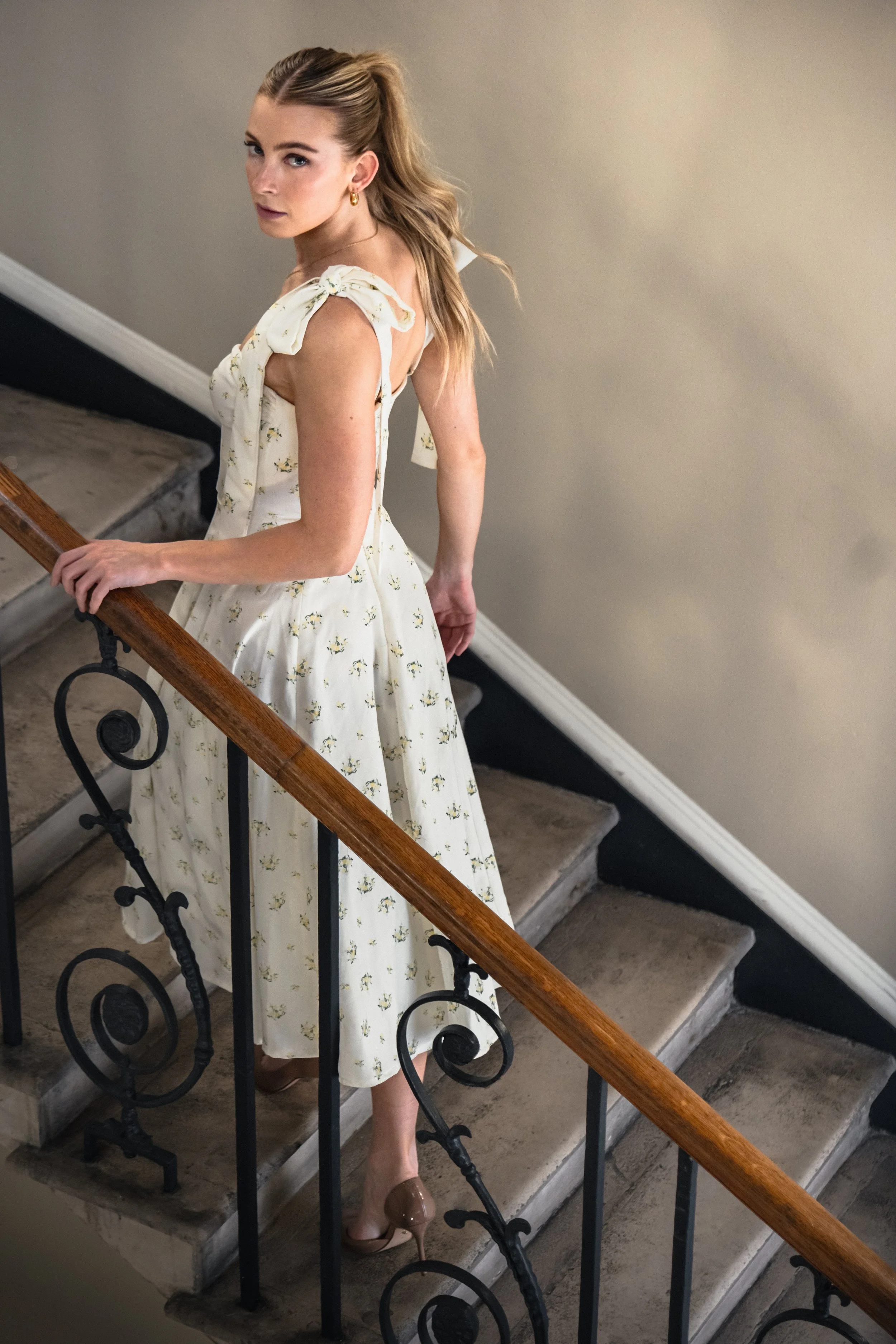 A young woman with blonde hair in a ponytail wearing a light-colored dress with small floral patterns, standing on a staircase and looking up at the camera.