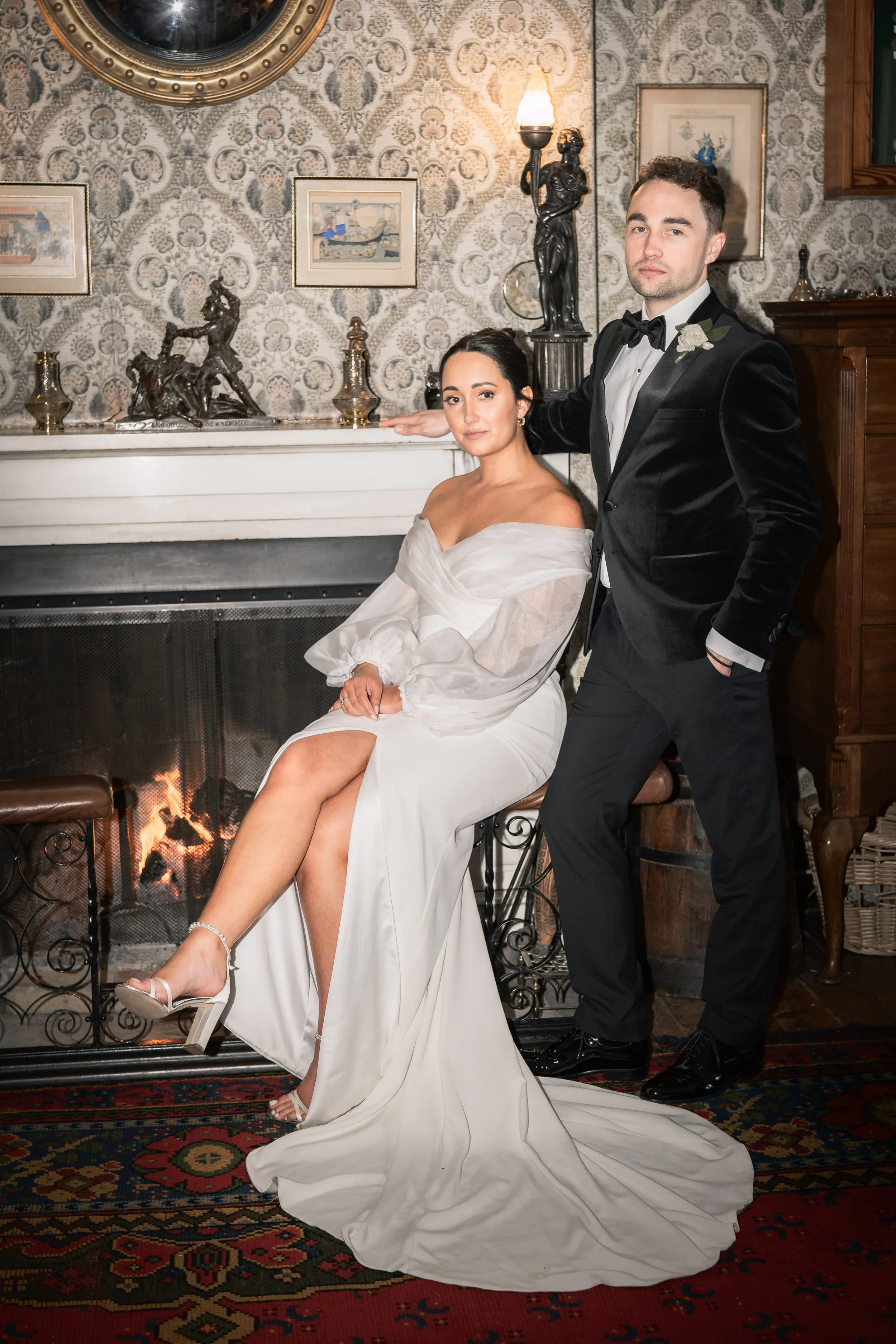 A bride and groom in formal attire posing indoors in front of a fireplace with ornate decorations and framed artwork. The bride is seated on a stool wearing a white off-shoulder gown, and the groom is standing beside her in a black tuxedo with a bow 