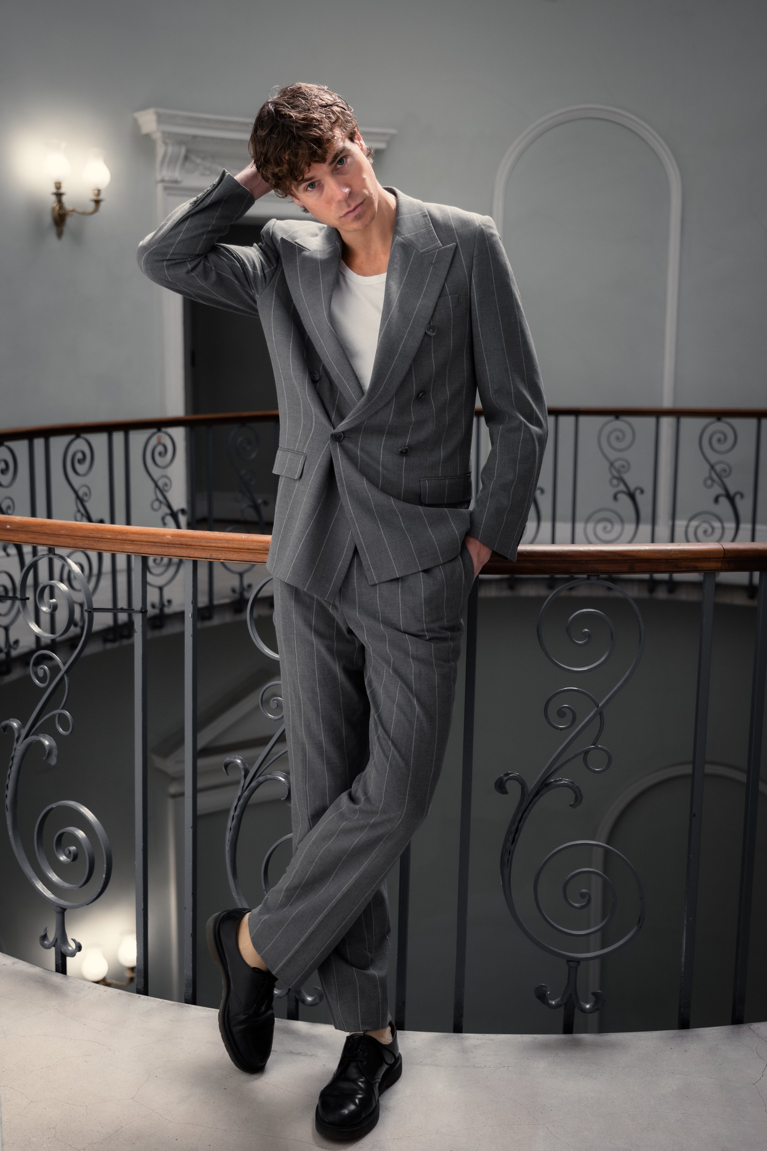 A young man in a gray pinstripe suit and white t-shirt leaning on a black ornate railing in a well-lit interior space.