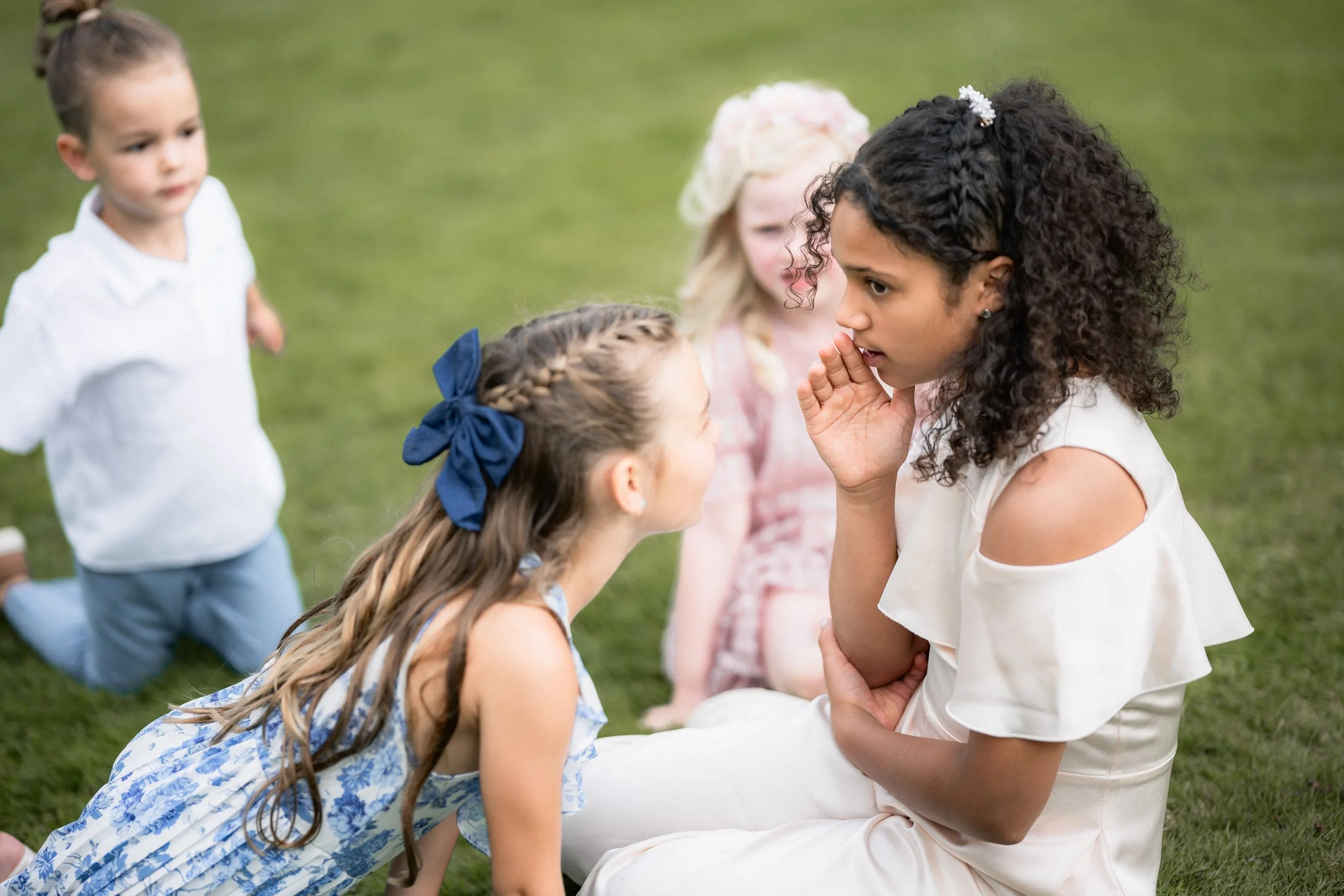 A woman with curly dark hair and tattoos on her arm is sitting on the grass, engaging in a serious conversation with a young girl who has braided hair and a blue bow. Two other children are nearby, one with a white shirt and bow in her hair, and anot