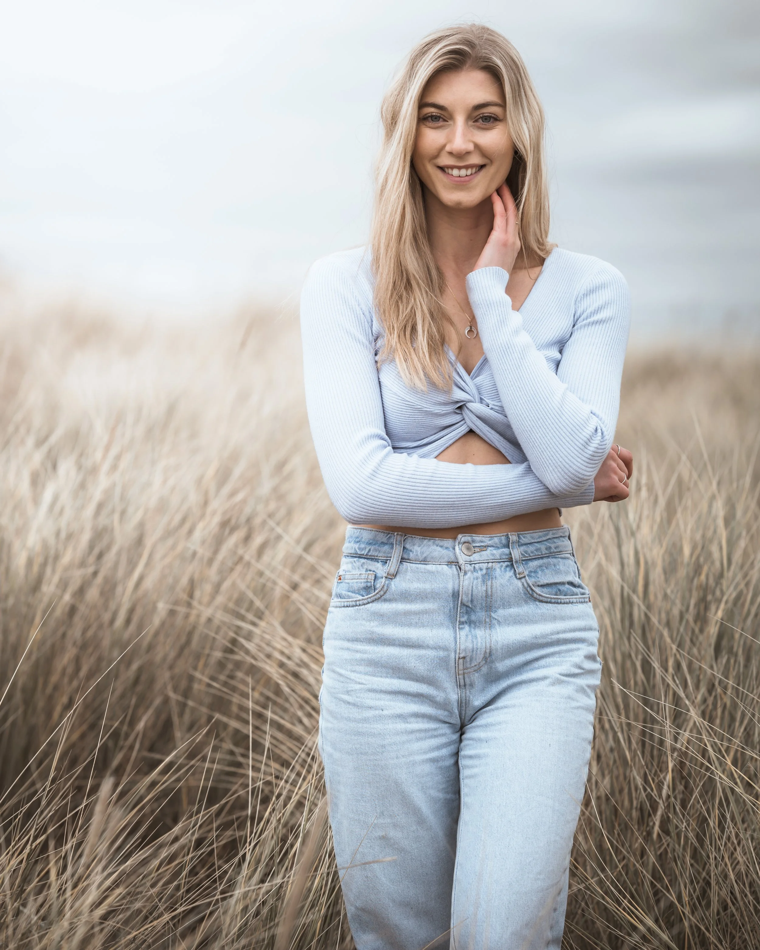 A young woman standing in a field of tall, dry grass, smiling at the camera, wearing a light blue long-sleeve top and high-waisted light wash jeans.